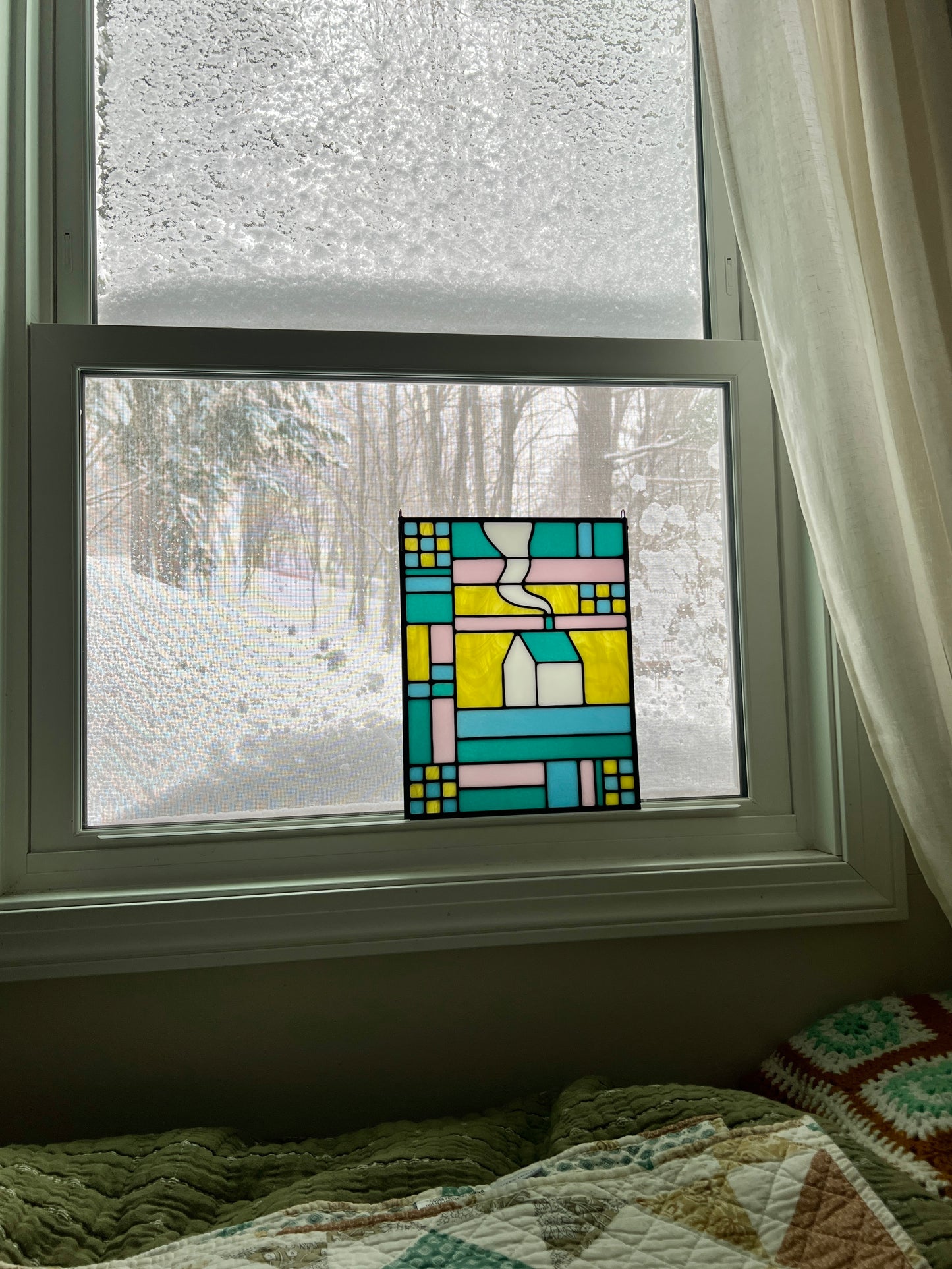 Stained glass textile inspired panel depicting a home in the center in shades of teal, pink, white, blue, and yellow, resting in a window with a snowy landscape outside