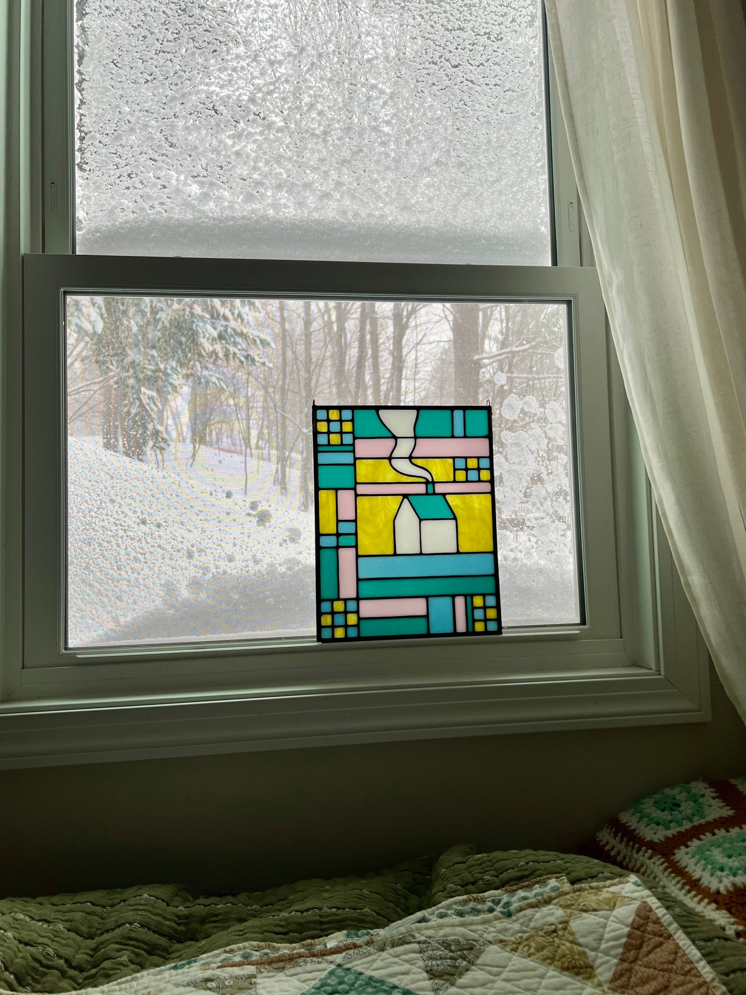 Stained glass textile inspired panel depicting a home in the center in shades of teal, pink, white, blue, and yellow, resting in a window with a snowy landscape outside