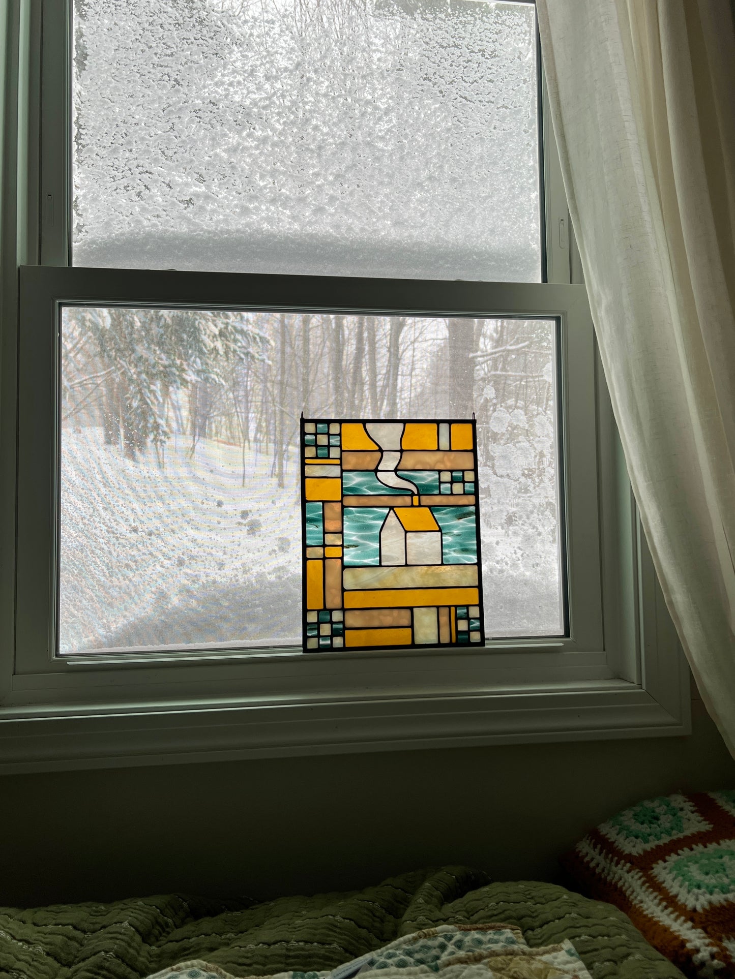 Stained glass textile inspired panel depicting a home in the center in shades of teal, amber, white, and beige, resting in a window with a snowy scene outside and cozy blankets in the foreground