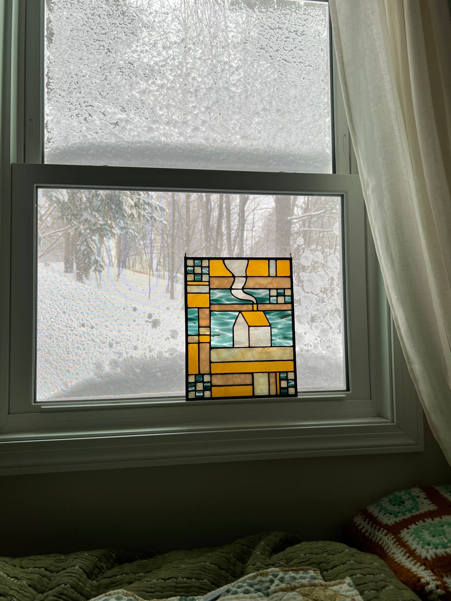 Stained glass textile inspired panel depicting a home in the center in shades of teal, amber, white, and beige, resting in a window with a snowy scene outside and cozy blankets in the foreground