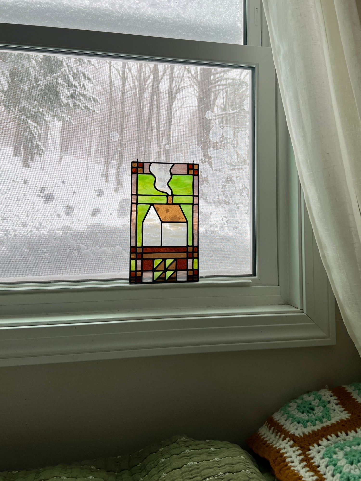 Stained glass textile inspired panel depicting a home in the center in shades of green, pink, white, and brown, resting in a window with a snowy scene outside.