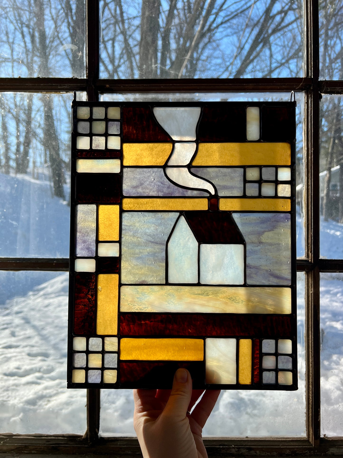 Stained glass textile inspired panel depicting a home in the center in shades of purple, white, brown, and amber, held by a hand in front of a brightly lit window with a snowy landscape outside