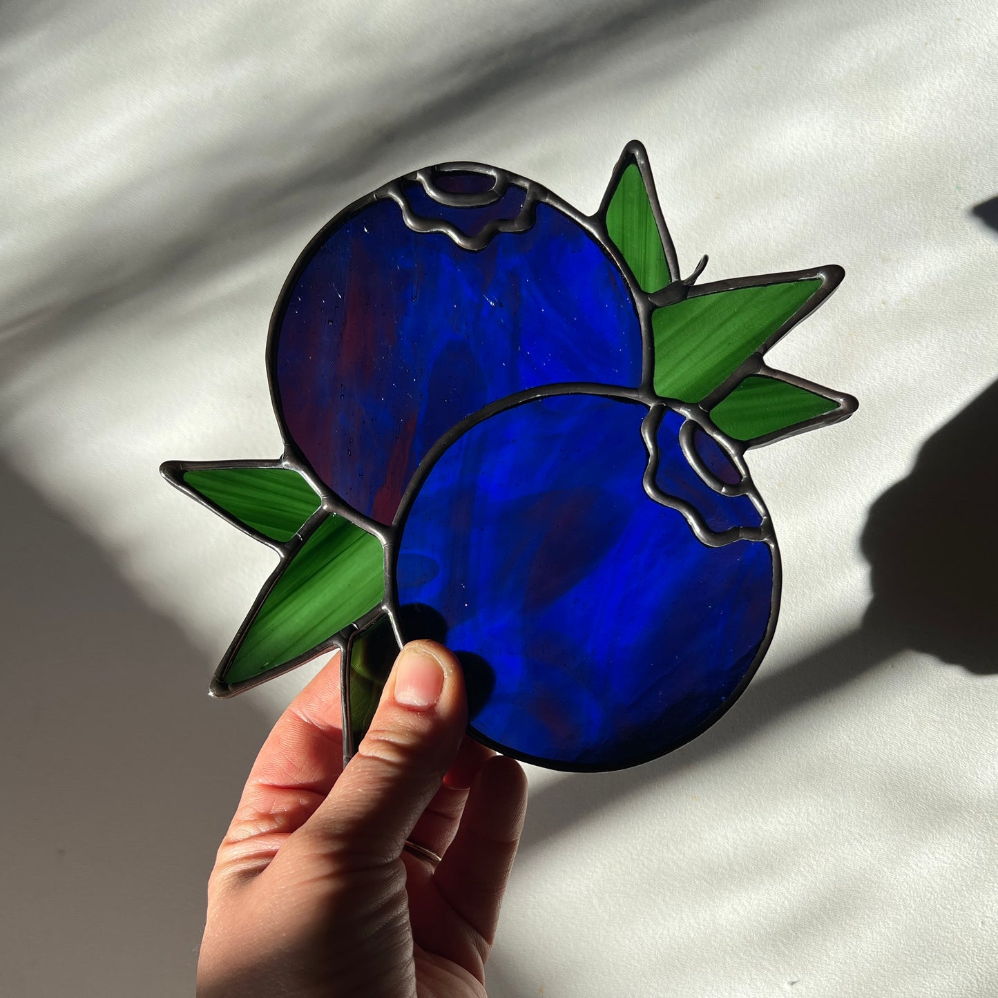 hand holding a wispy blue stained glass suncatcher depicting a small bunch of blueberries with leaves