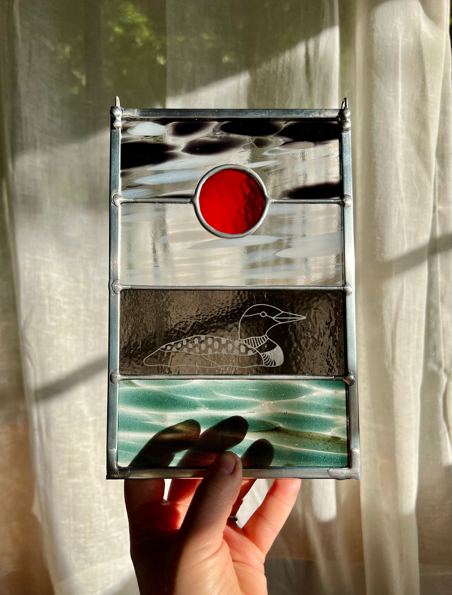 a hand holding a stained glass panel with black, white, clear, teal, and red glass with a loon etched on the surface