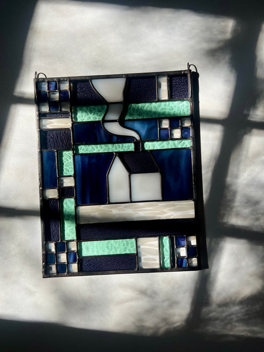 Stained glass panel with geometric textile inspired design with a cozy home depicted in the center, laying on a white black ground with bright sunlight across it