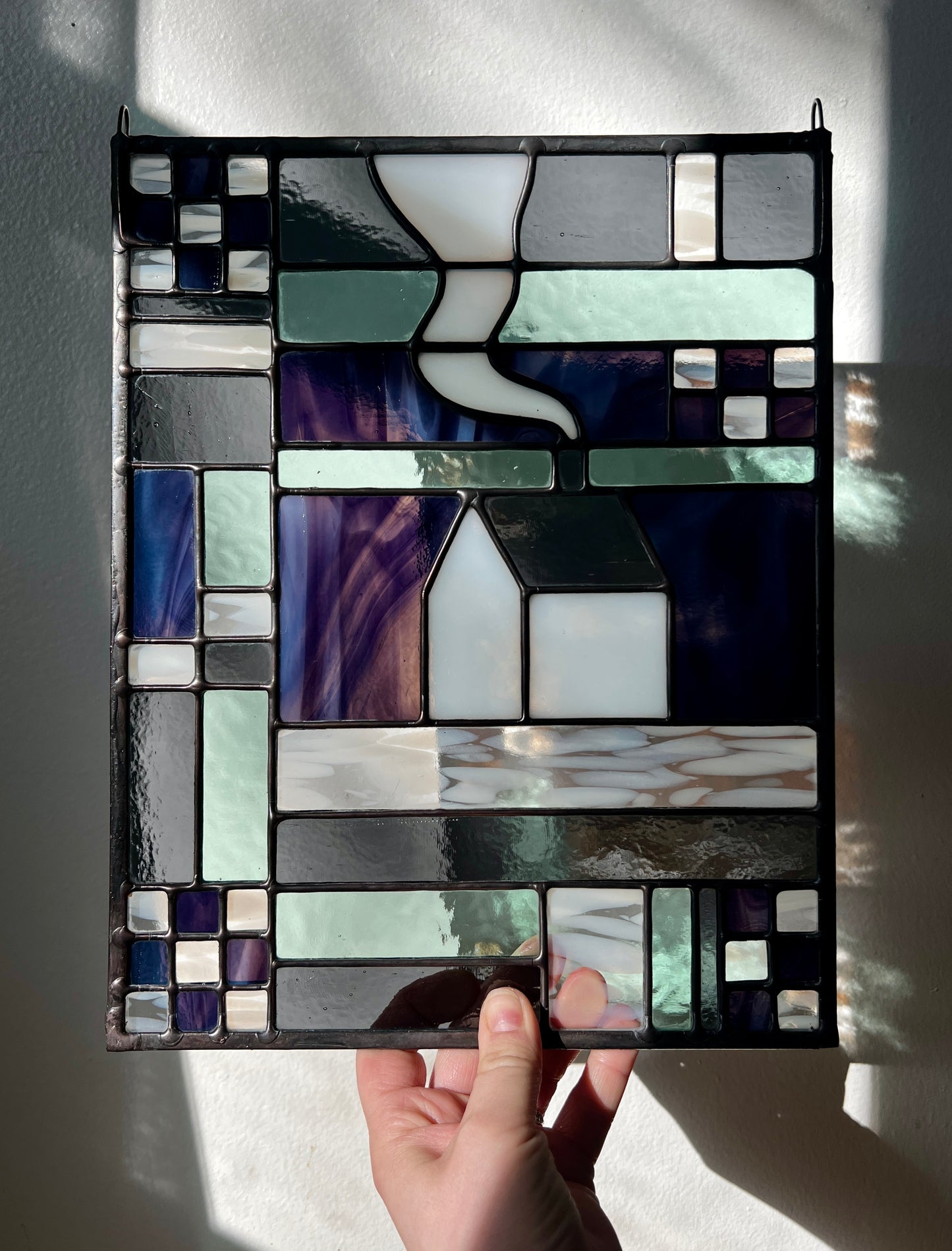 Stained glass panel with geometric textile inspired design with a cozy home depicted in the center, held by a hand against a white background.