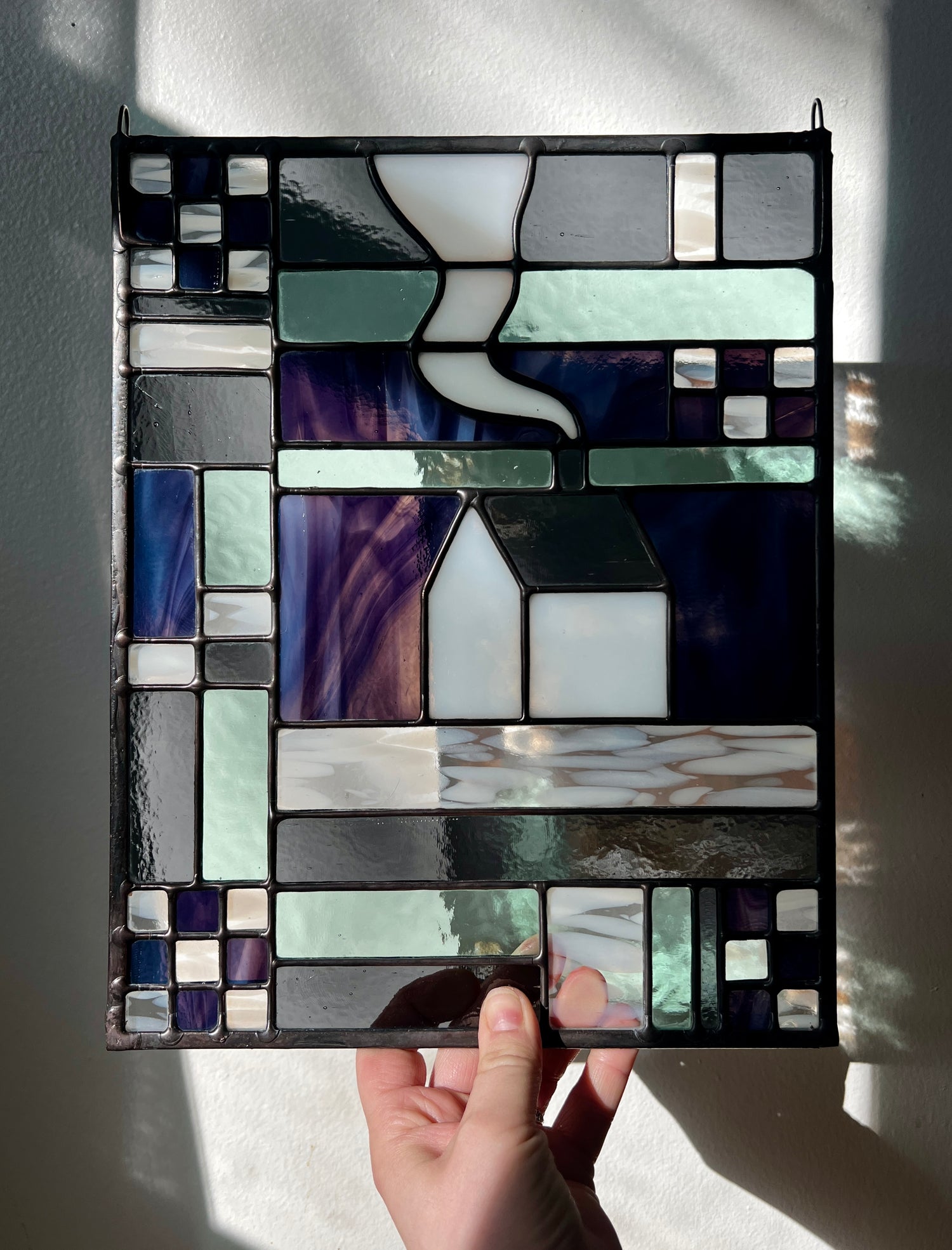 Stained glass panel with geometric textile inspired design with a cozy home depicted in the center, held by a hand against a white background.