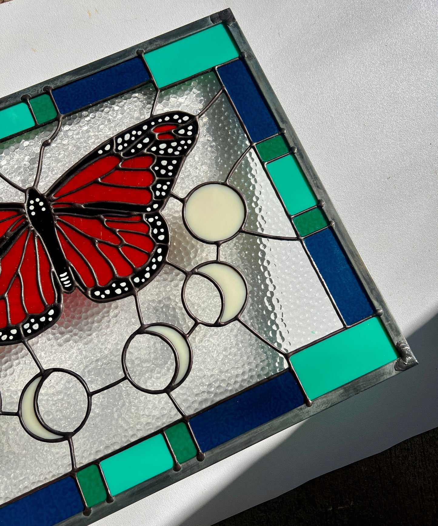 A large stained glass panel with a thick blue and green geometric border and a monarch butterfly and moon phases in the center, with textured clear glass in the background, photographed at an angle against a white background.