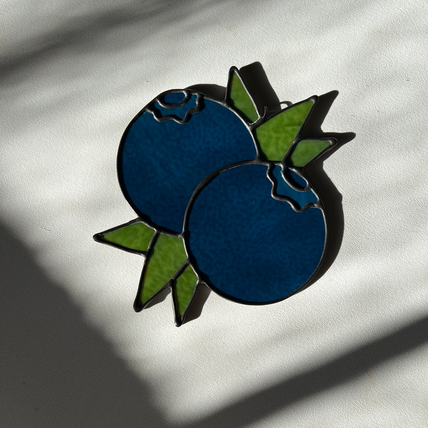 a dark blue stained glass suncatcher depicting a small bunch of blueberries with leaves resting on a white surface
