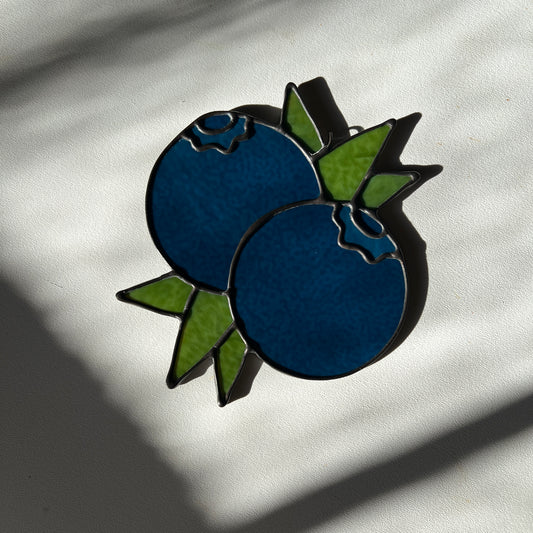 a dark blue stained glass suncatcher depicting a small bunch of blueberries with leaves resting on a white surface