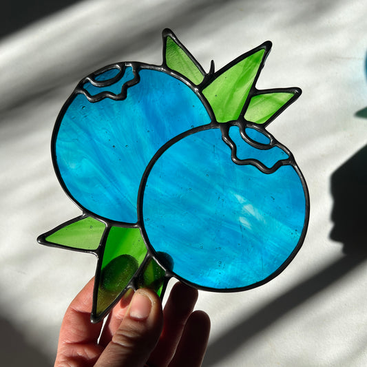 a hand holding a wispy blue suncatcher depicting a small bunch of blueberries with leaves
