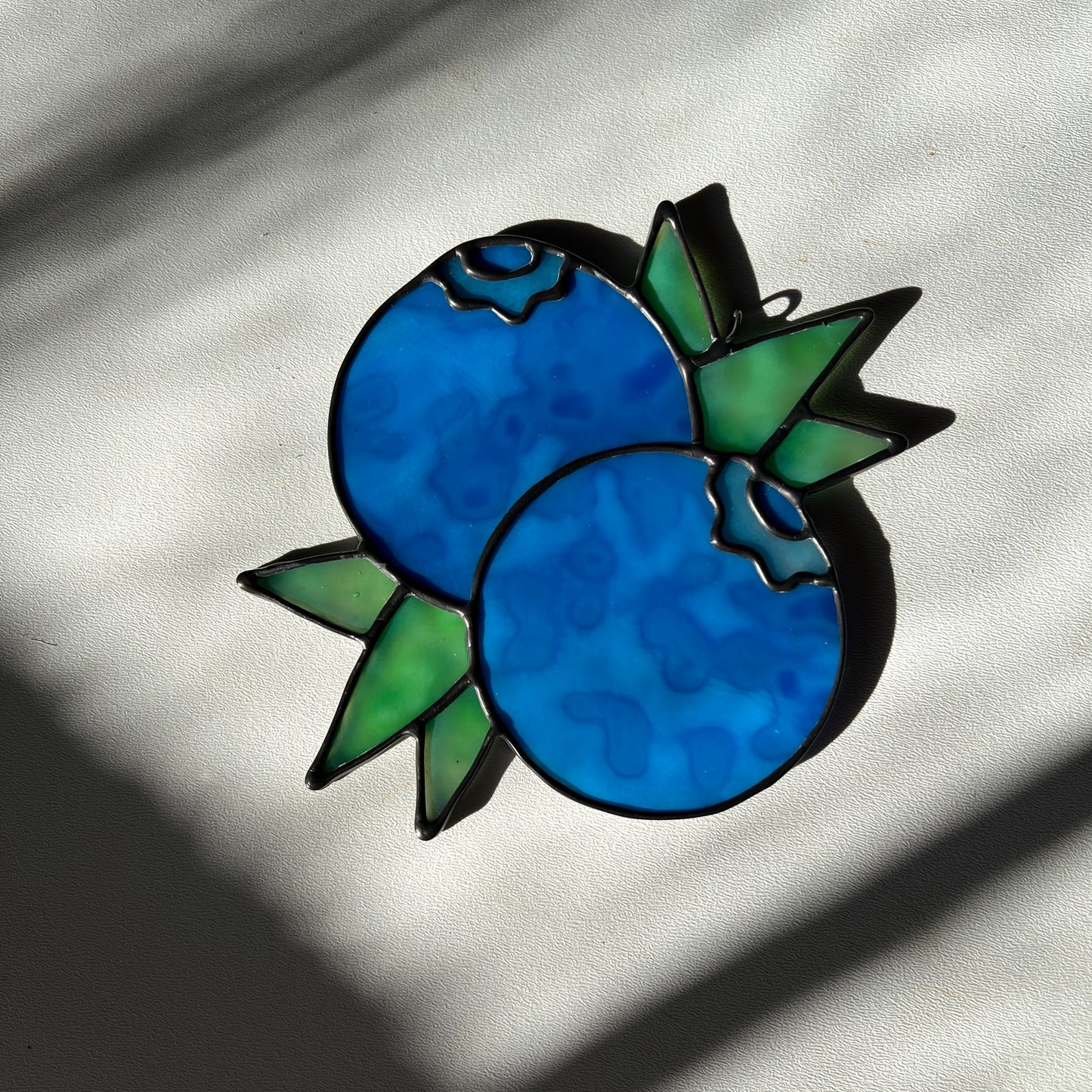 a mottled blue stained glass suncatcher depicting a small bunch of blueberries with leaves resting on a white surface