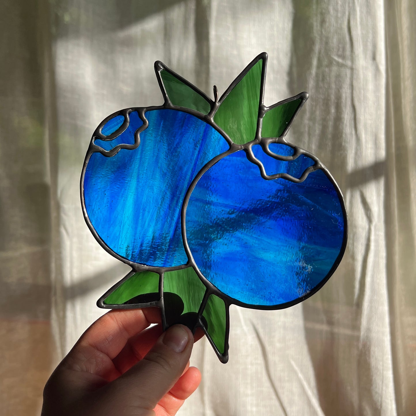 hand holding a wispy blue stained glass suncatcher depicting a small bunch of blueberries with leaves in front of a window