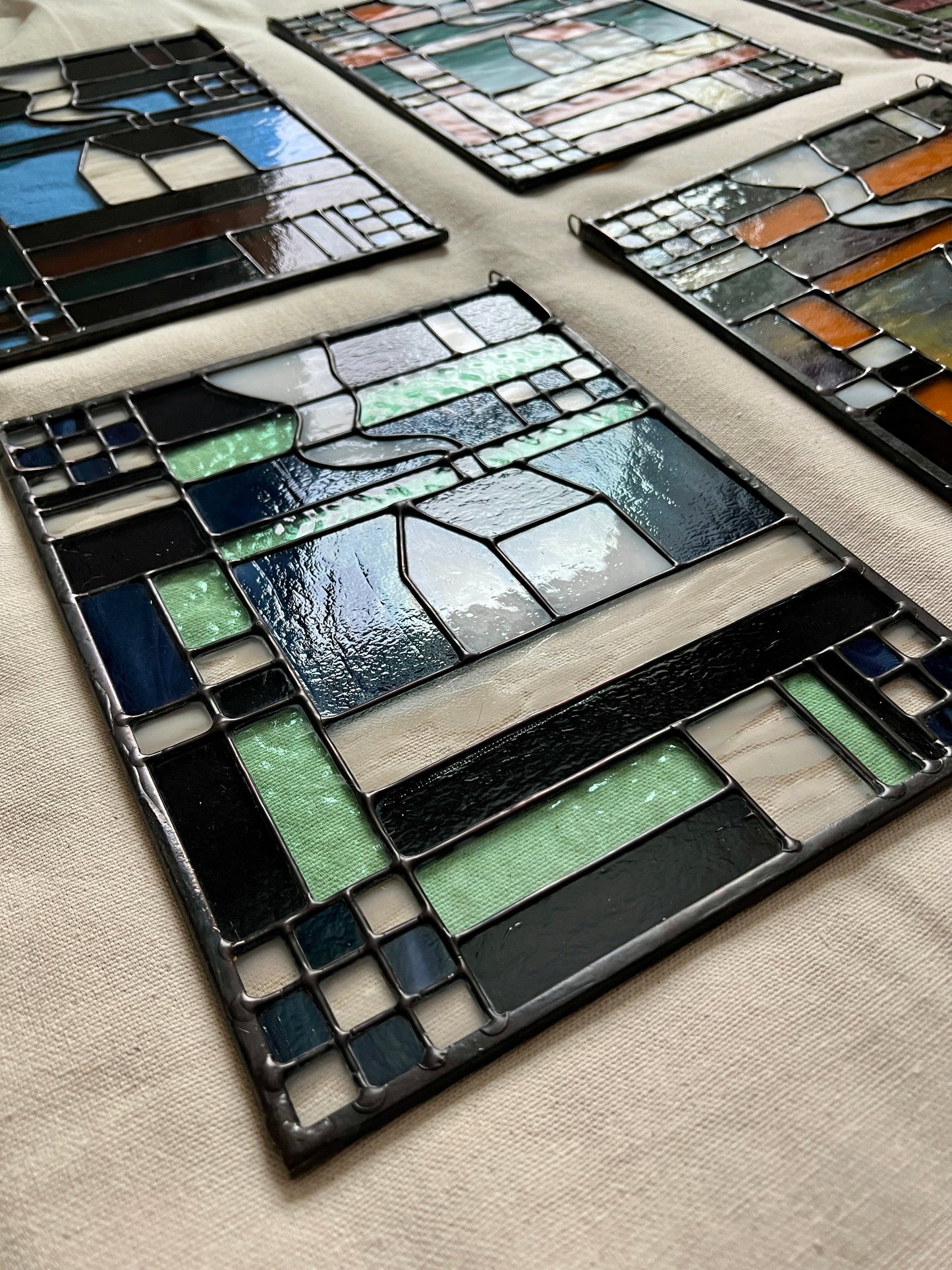 Close up photo of a stained glass panel with geometric textile inspired design with a cozy home depicted in the center, laying on a neutral fabric. The photo is taken at an angle to show smooth solder lines.