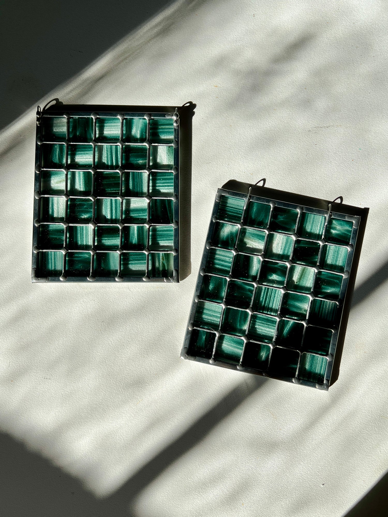 two stained glass suncatchers depicting streaky and mottled sparkly clear and teal glass arranged in a grid pattern, resting on a white surface