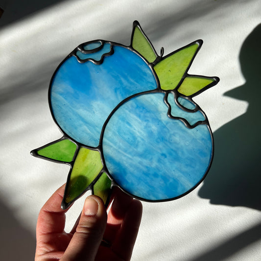 hand holding a wispy light blue stained glass suncatcher depicting a small bunch of blueberries with leaves