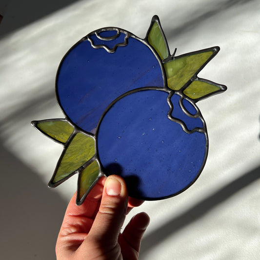 hand holding a blue stained glass suncatcher depicting a small bunch of blueberries with leaves