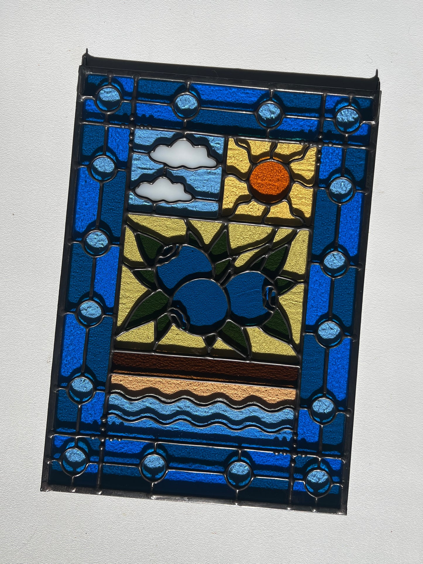 stained glass panel with a simple compartment style design featuring clouds, sun, water, and a small bunch of blueberries in the middle with a blue border