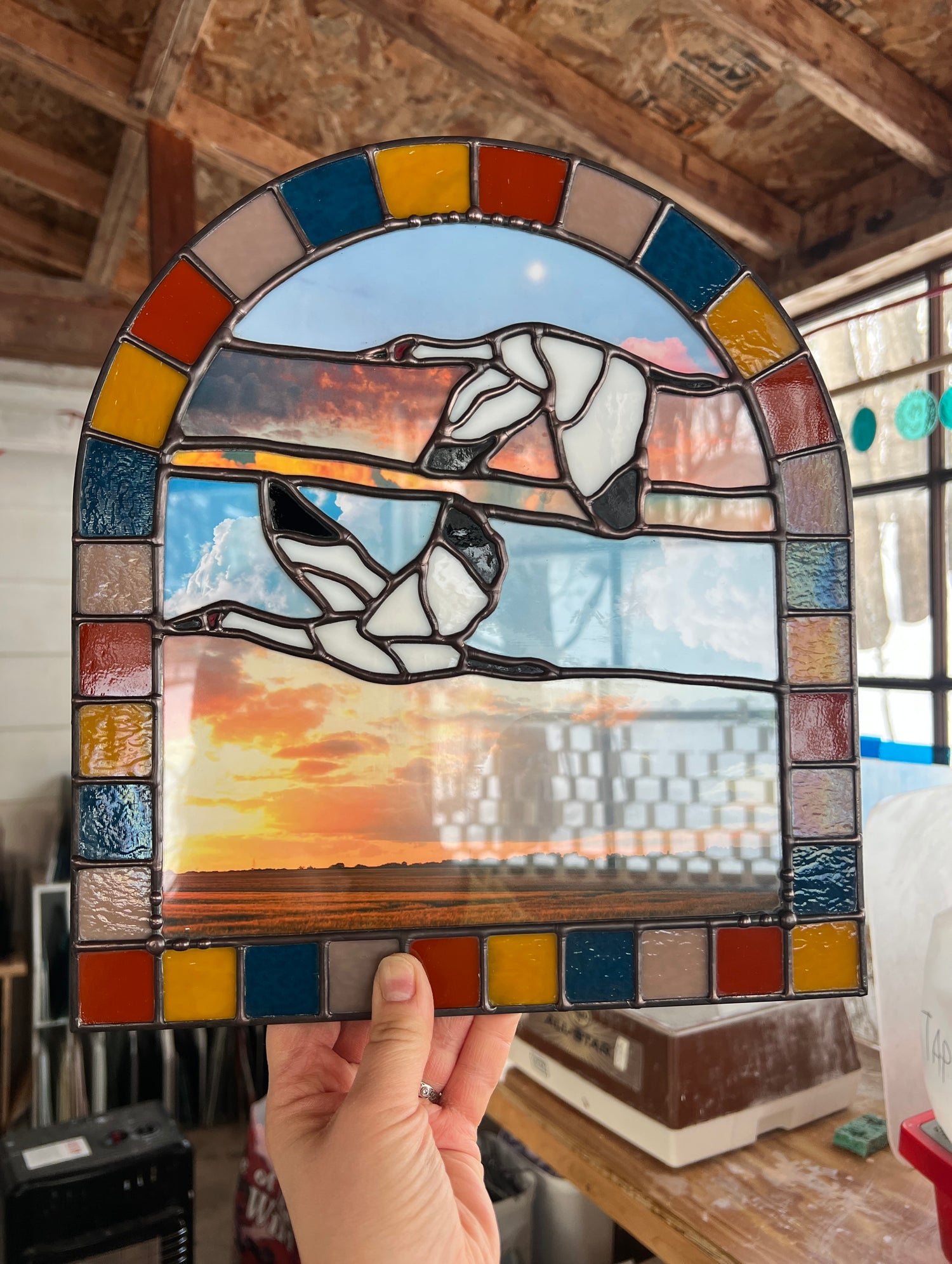 Stained glass panel with a landscape design created by collaged photographs and two cranes flying, held by a person indoors.