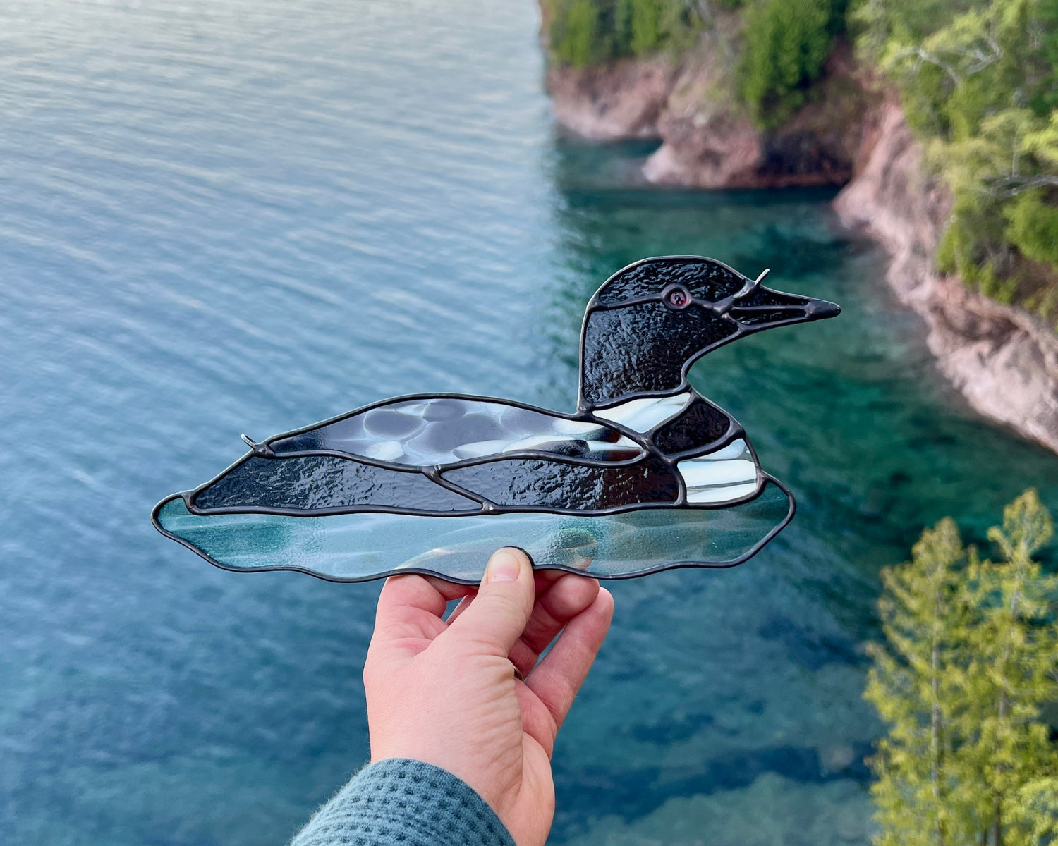 Hand holding a stained glass loon ornament over a cliffside overlooking turquoise colored lake superior and opposing cliff shoreline.
