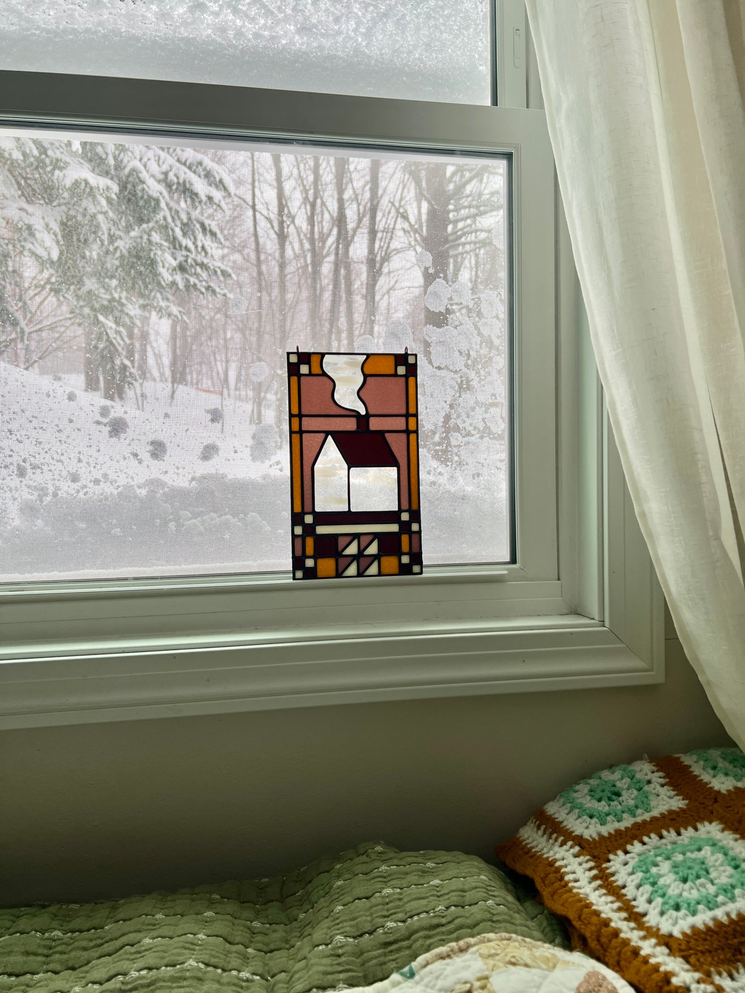 Stained glass textile inspired panel depicting a home in the center in shades of purple, white, brown, and bronze, resting in a window with a snowy scene outside and cozy blankets in the foreground