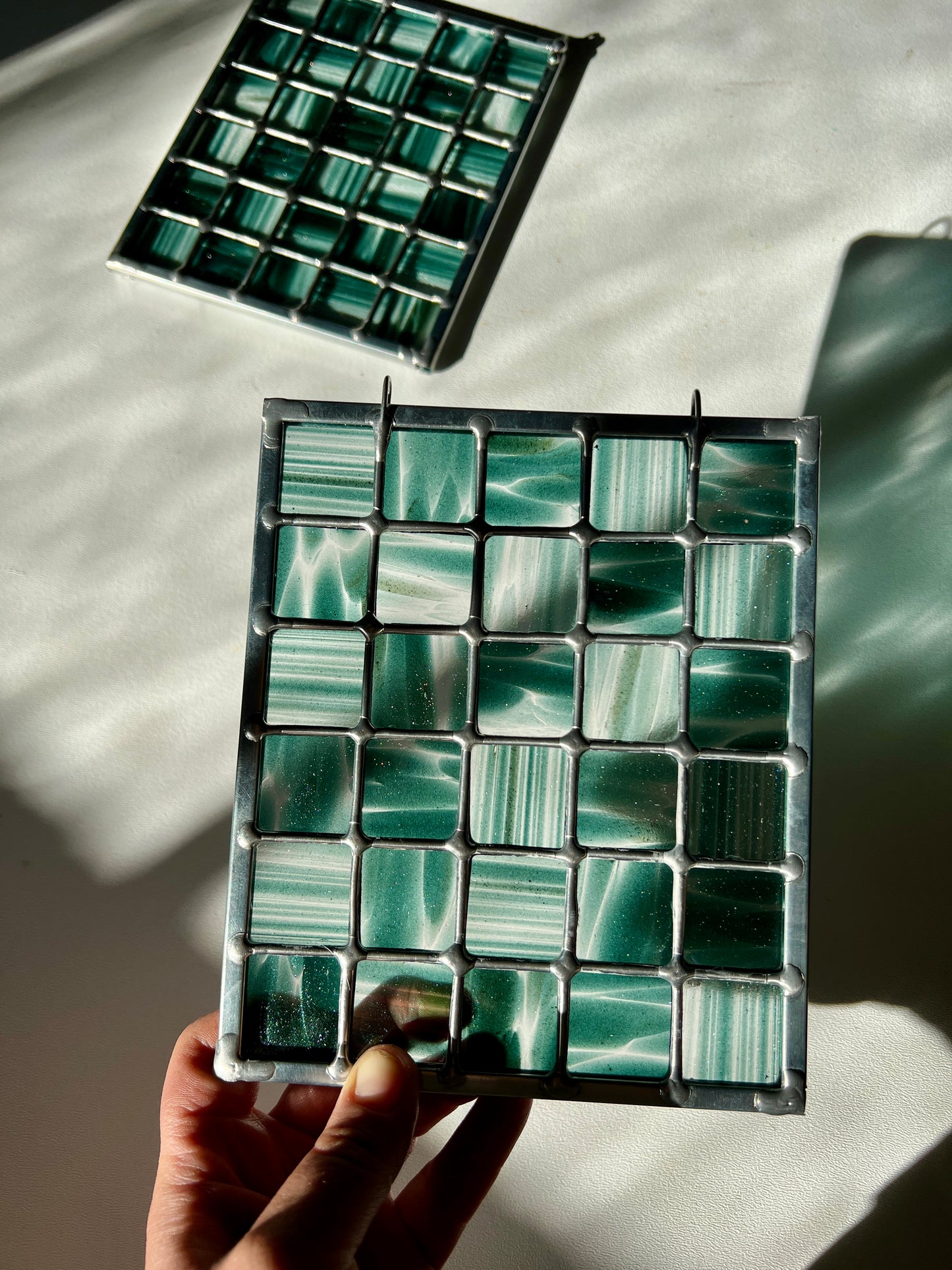 hand holding a stained glass suncatcher depicting streaky and mottled sparkly clear and teal glass arranged in a grid pattern, held above a white surface with sunlight casting a partial refraction, with a second similar suncatcher in the background