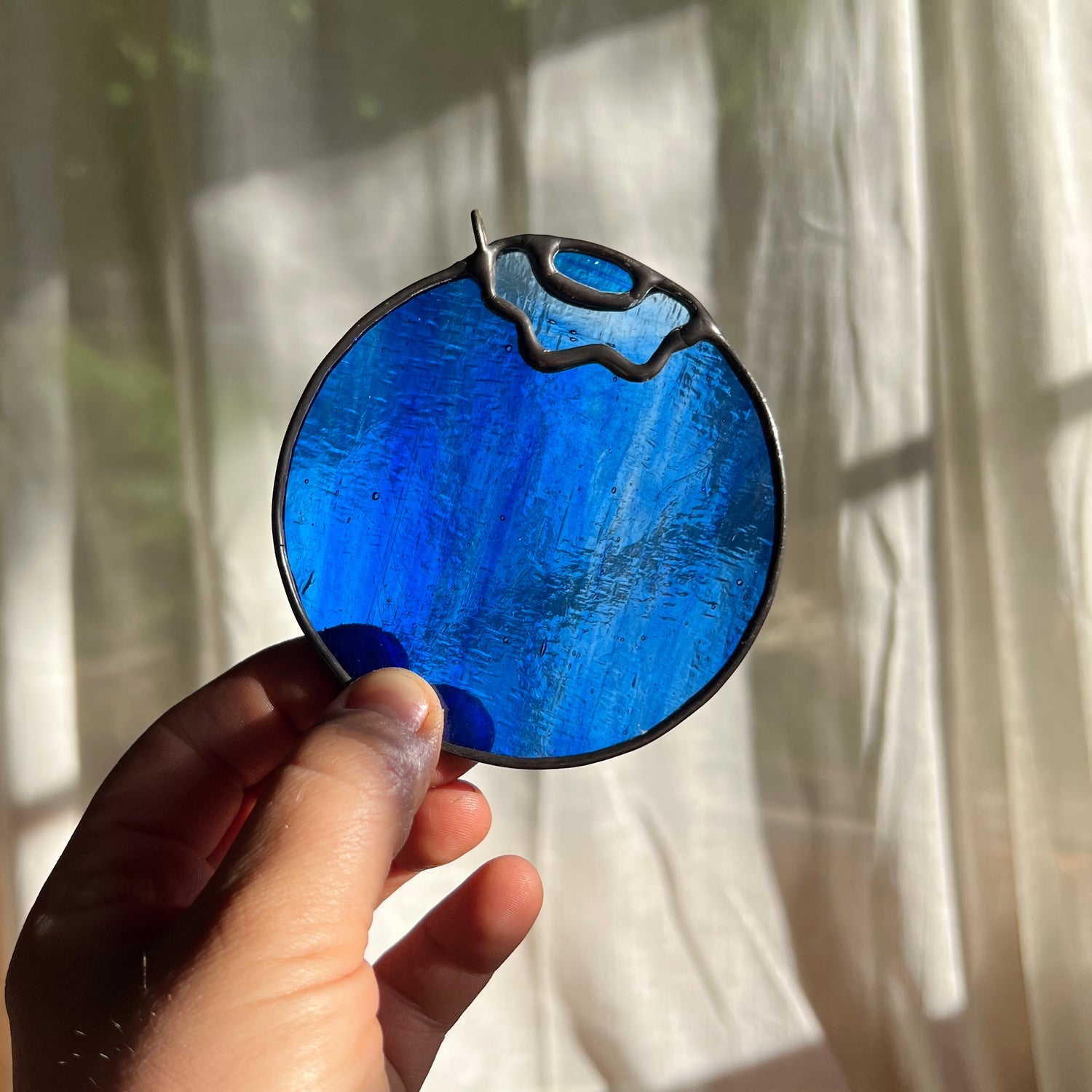 hand holding a blue wispy stained glass blueberry in front of window