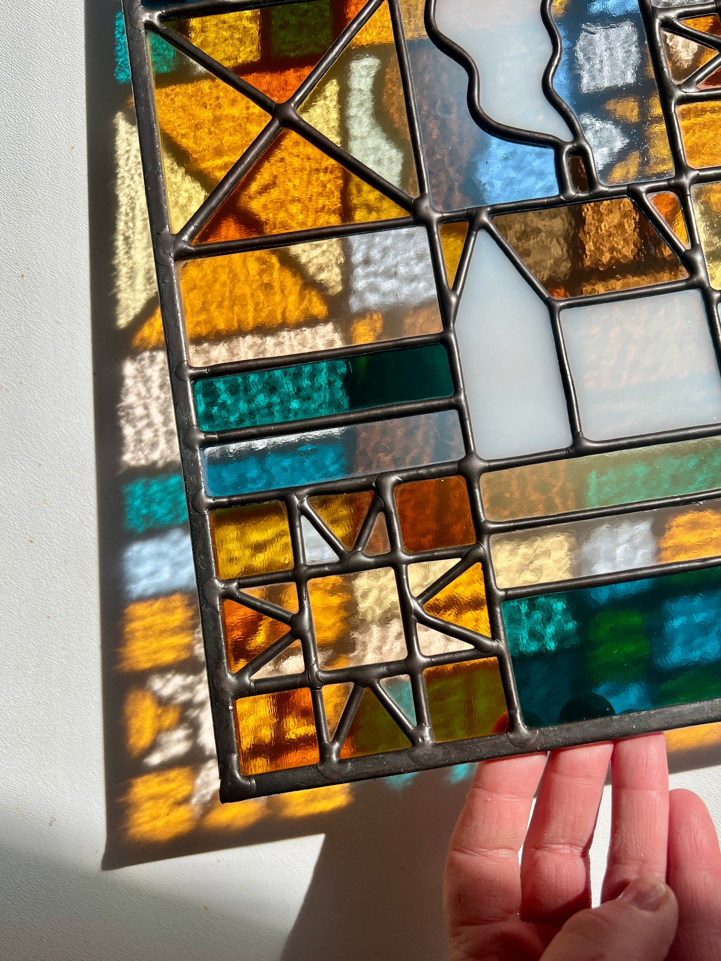 Close up of a stained glass textile inspired panel depicting a home in the center in shades of teal, pink, white, blue, and yellow, held by a hand against a white background with sunlight casting colorful refractions