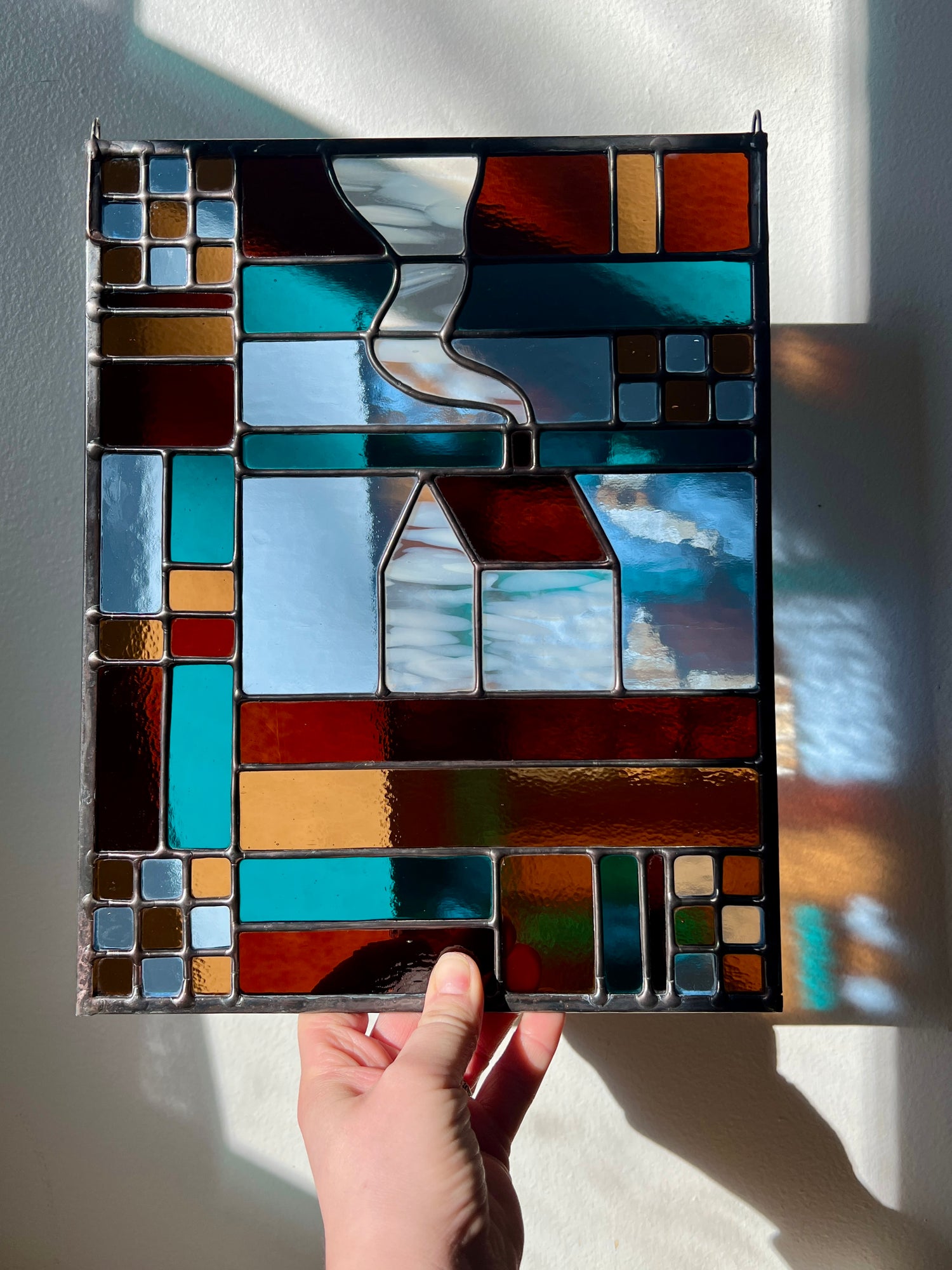 Stained glass textile inspired panel depicting a home in the center in shades of teal, brown, and blue, held by a hand against a white wall with sunlight casting refractions