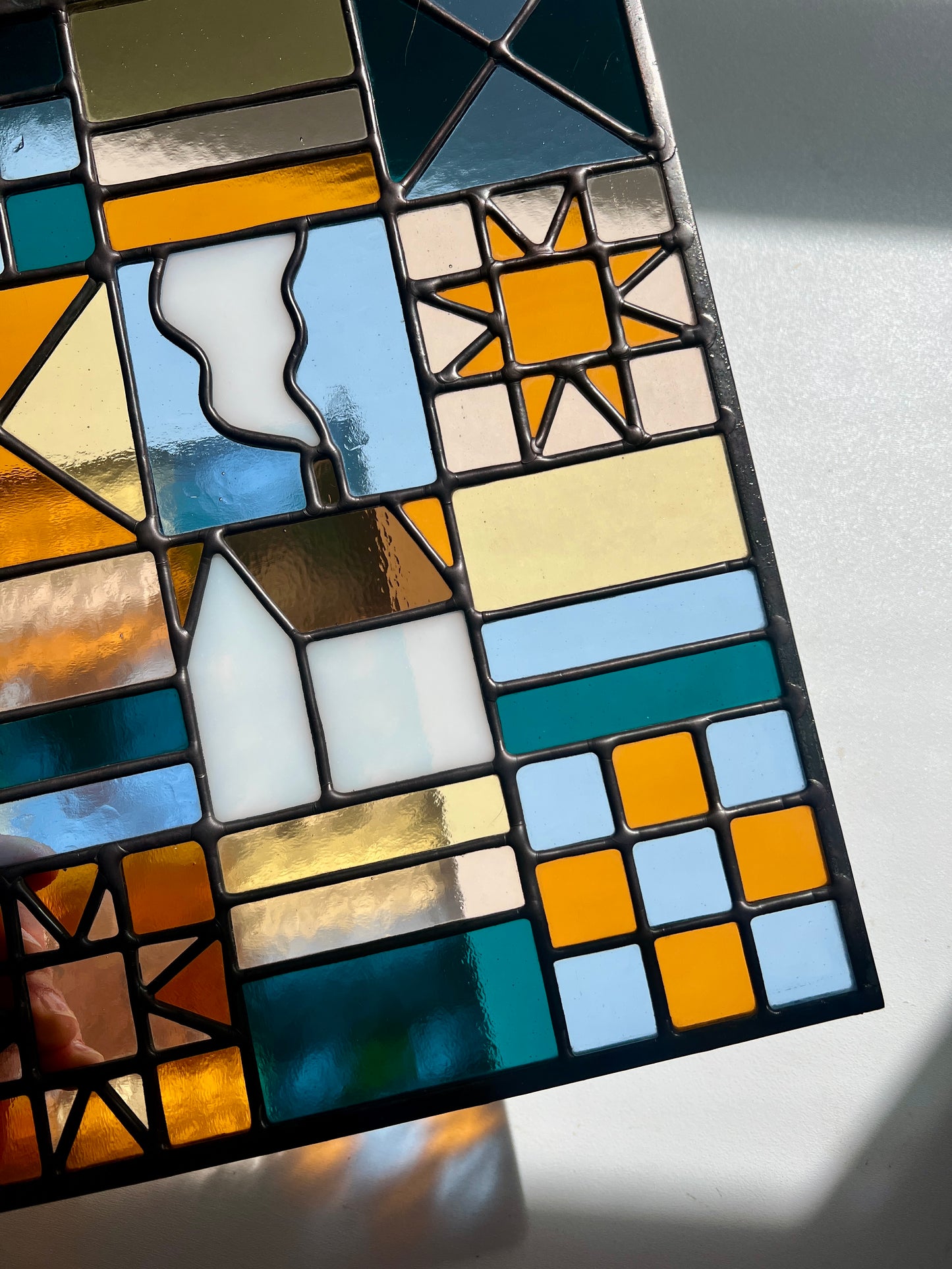 Close up of a textile inspired stained glass panel depicting a home in the center in shades of teal, pink, white, blue, and yellow,  held above a white background