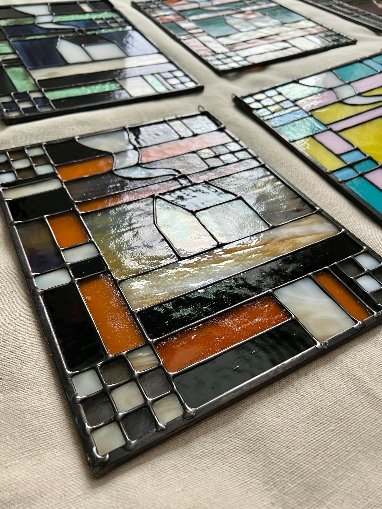 Stained glass textile inspired panel depicting a home in the center in shades of purple, white, brown, and amber, photographed at an angle to highlight smooth solder lines