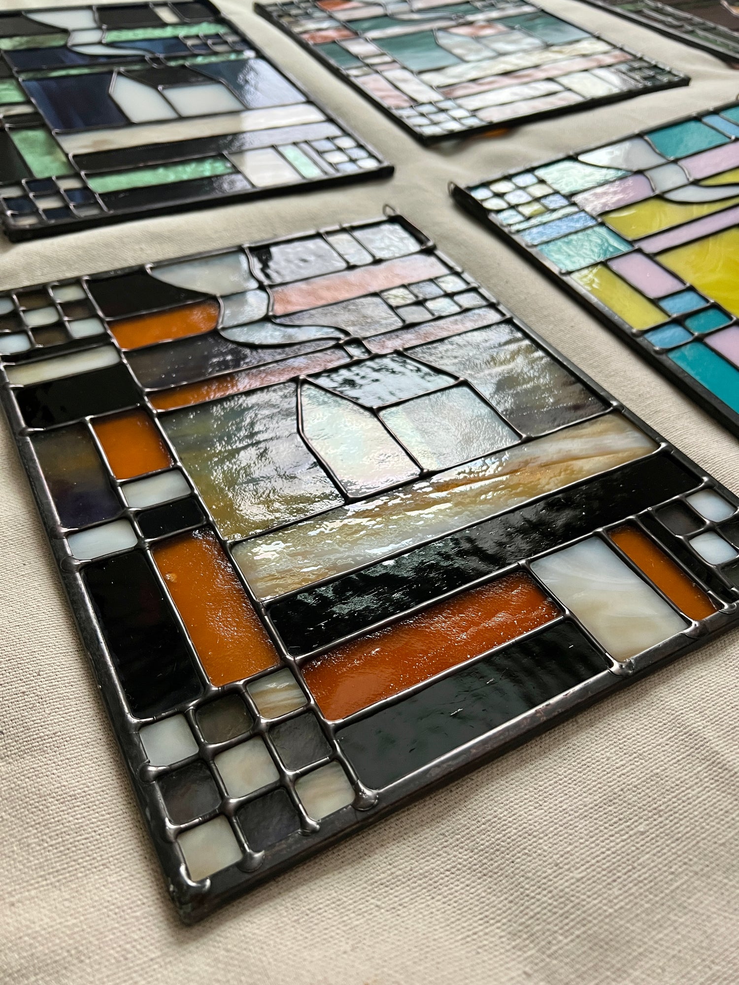 Stained glass textile inspired panel depicting a home in the center in shades of purple, white, brown, and amber, photographed at an angle to highlight smooth solder lines