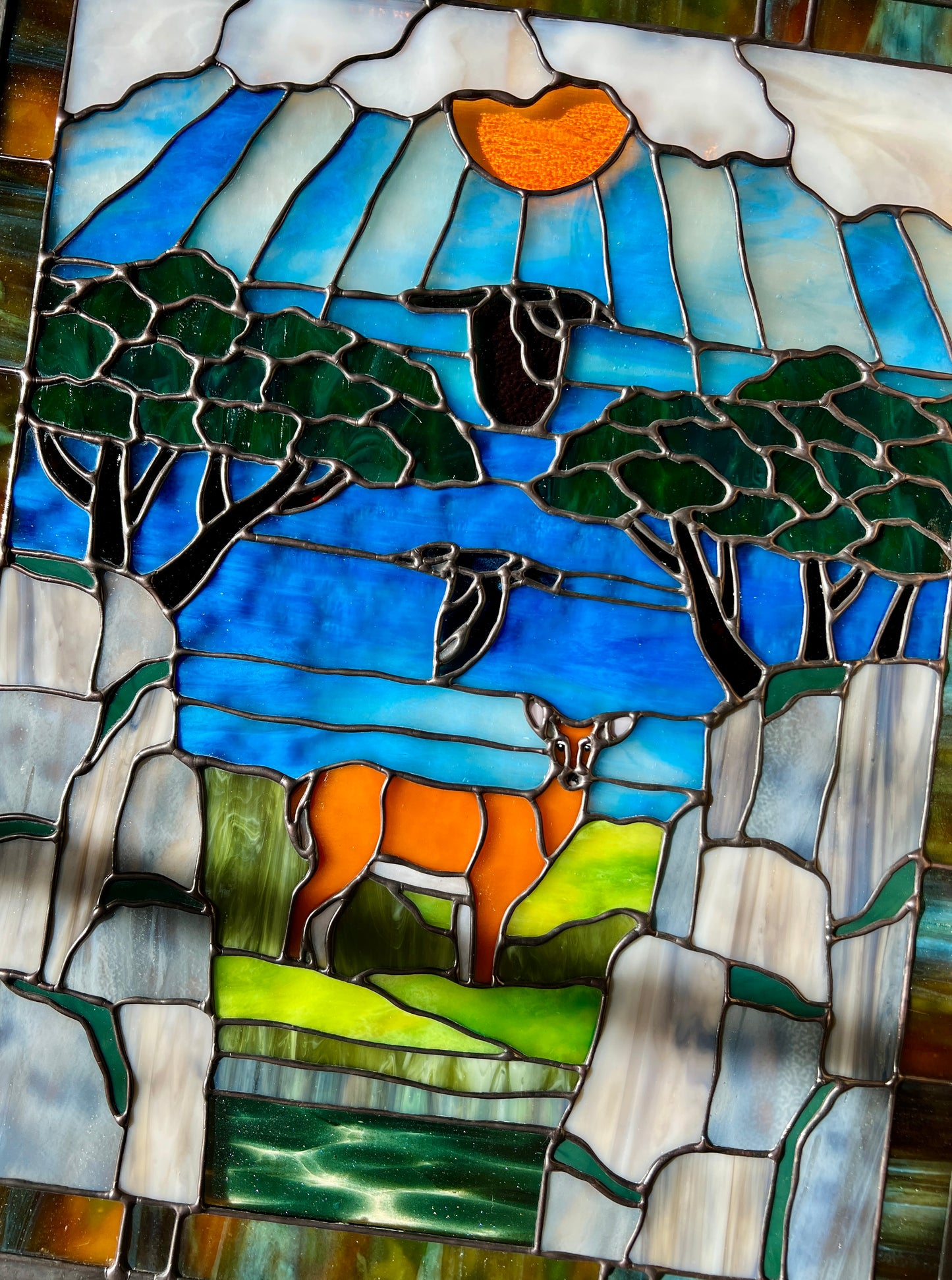 large stained glass panel depicting mossy, rocky cliffs and trees, with a deer, a kingfisher, and a raven standing amongst a river, greenery, and bright blue skies with some clouds and a bright yellow orange sun.