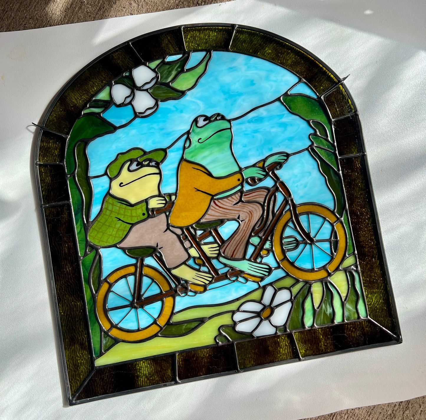 stained glass panel depicting frog and toad riding a bicycle resting on a white surface