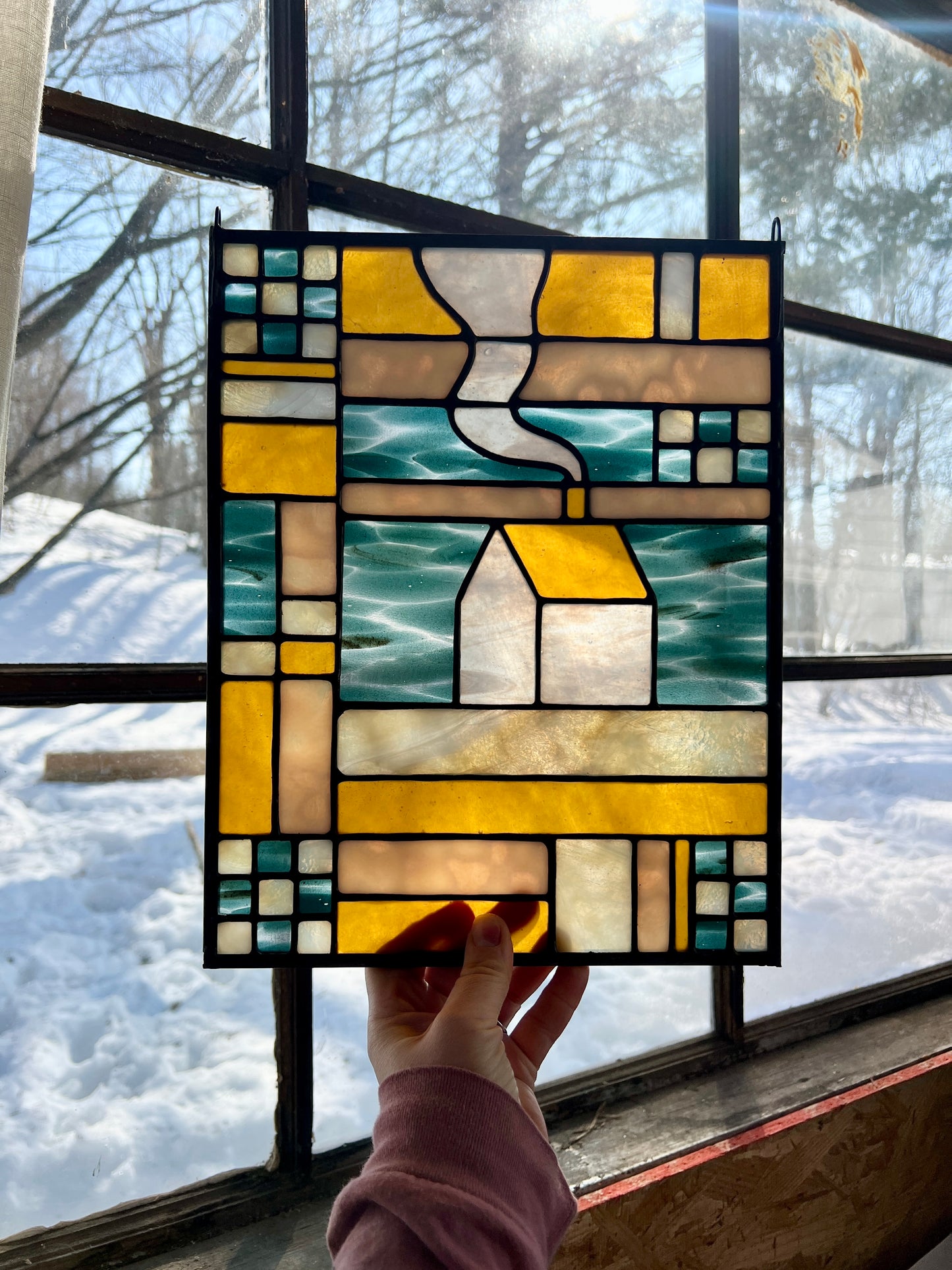 Stained glass textile inspired panel depicting a home in the center in shades of teal, amber, white, and beige, held by a hand against a large gently backlit window with a snowy scene outside