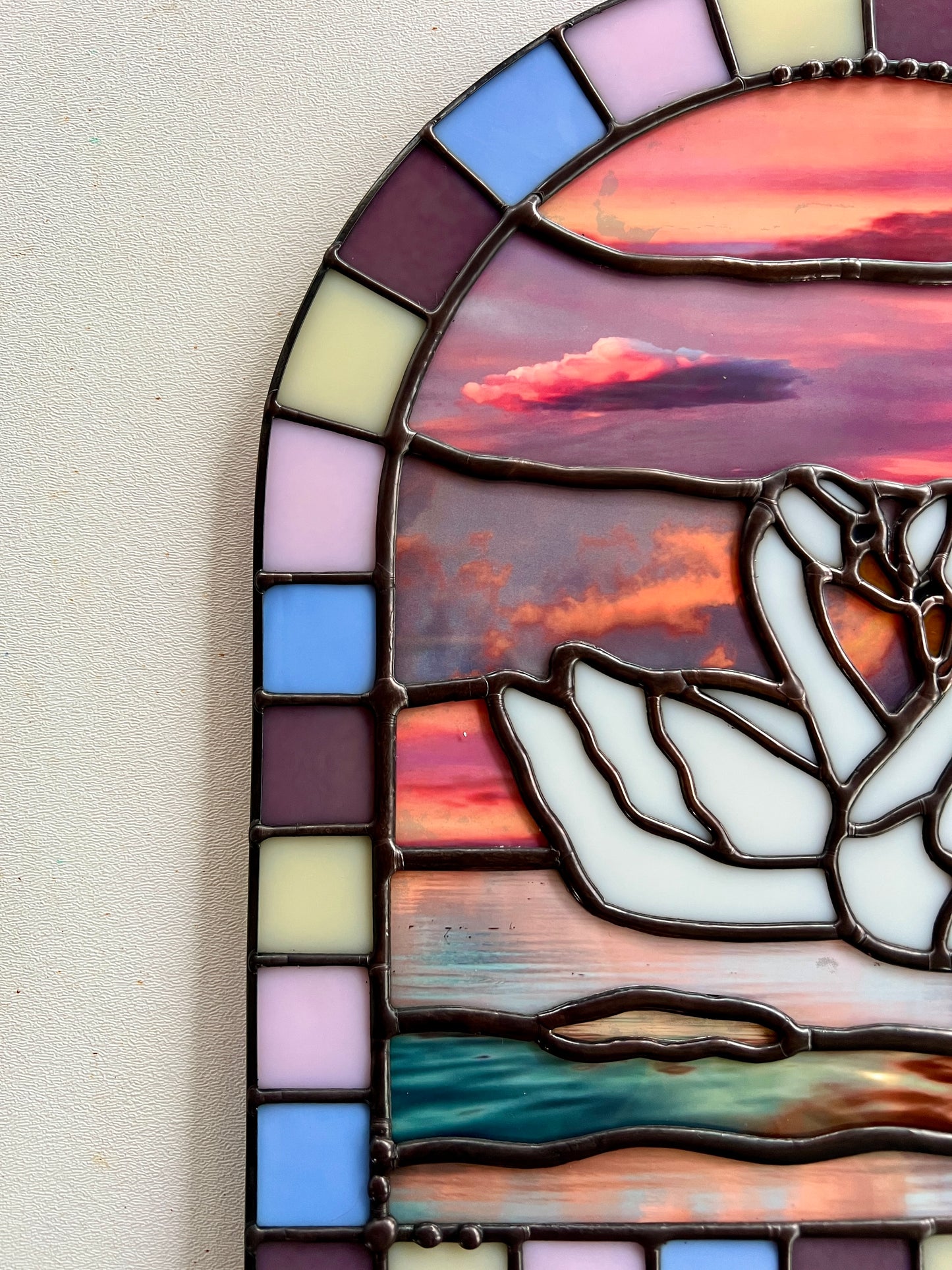 Close up of a stained glass panel with a colorful pastel arch border and two white swans in the center. Collaged photographs are used to create a sunset background.