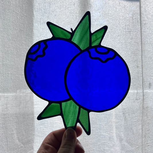 A hand holding a stained glass suncatcher depicting two blueberries and some leaves against a gently backlit white background.