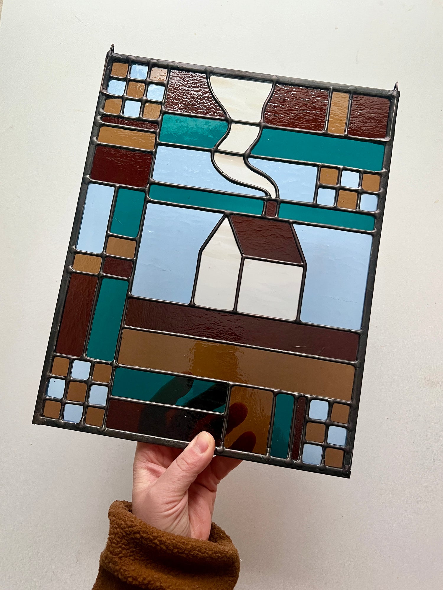 Stained glass textile inspired panel depicting a home in the center in shades of teal, brown, and blue, held by a hand against a white backdrop