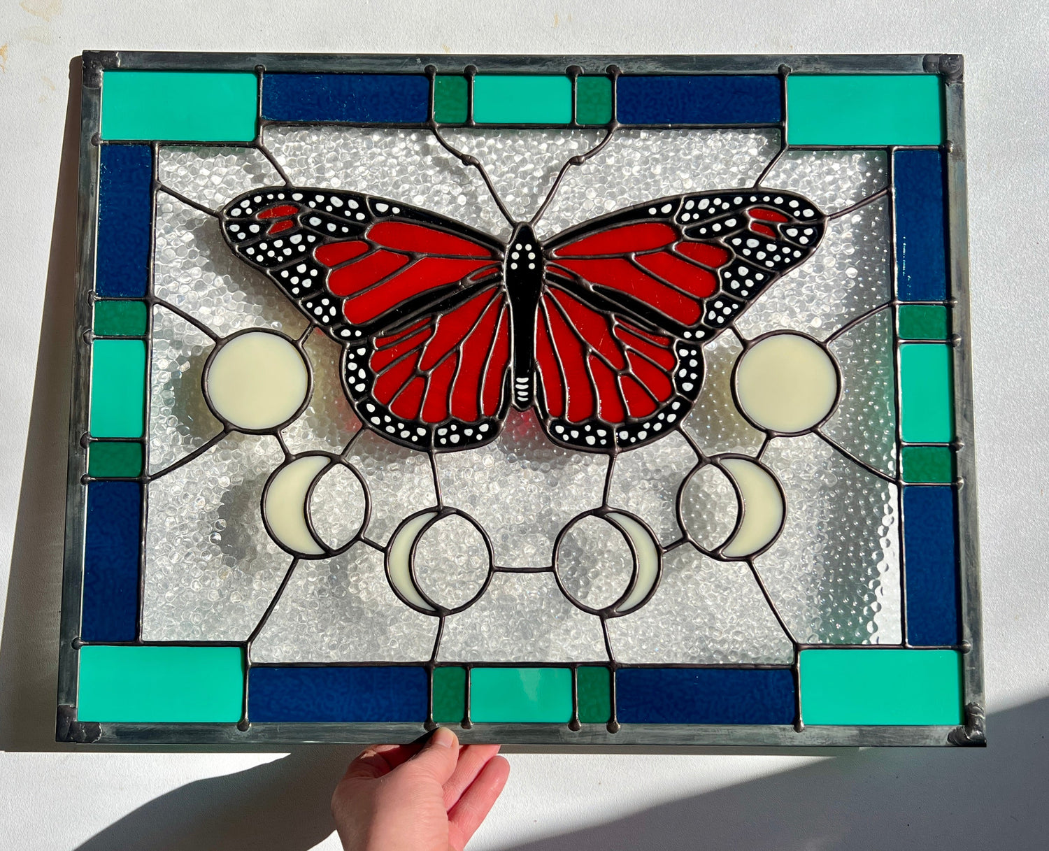A rectangular stained glass panel with a thick blue and green geometric border and a monarch butterfly and moon phases in the center, with textured clear glass in the background. The piece is held by a hand against a white background, with bright sunlight flowing across it.