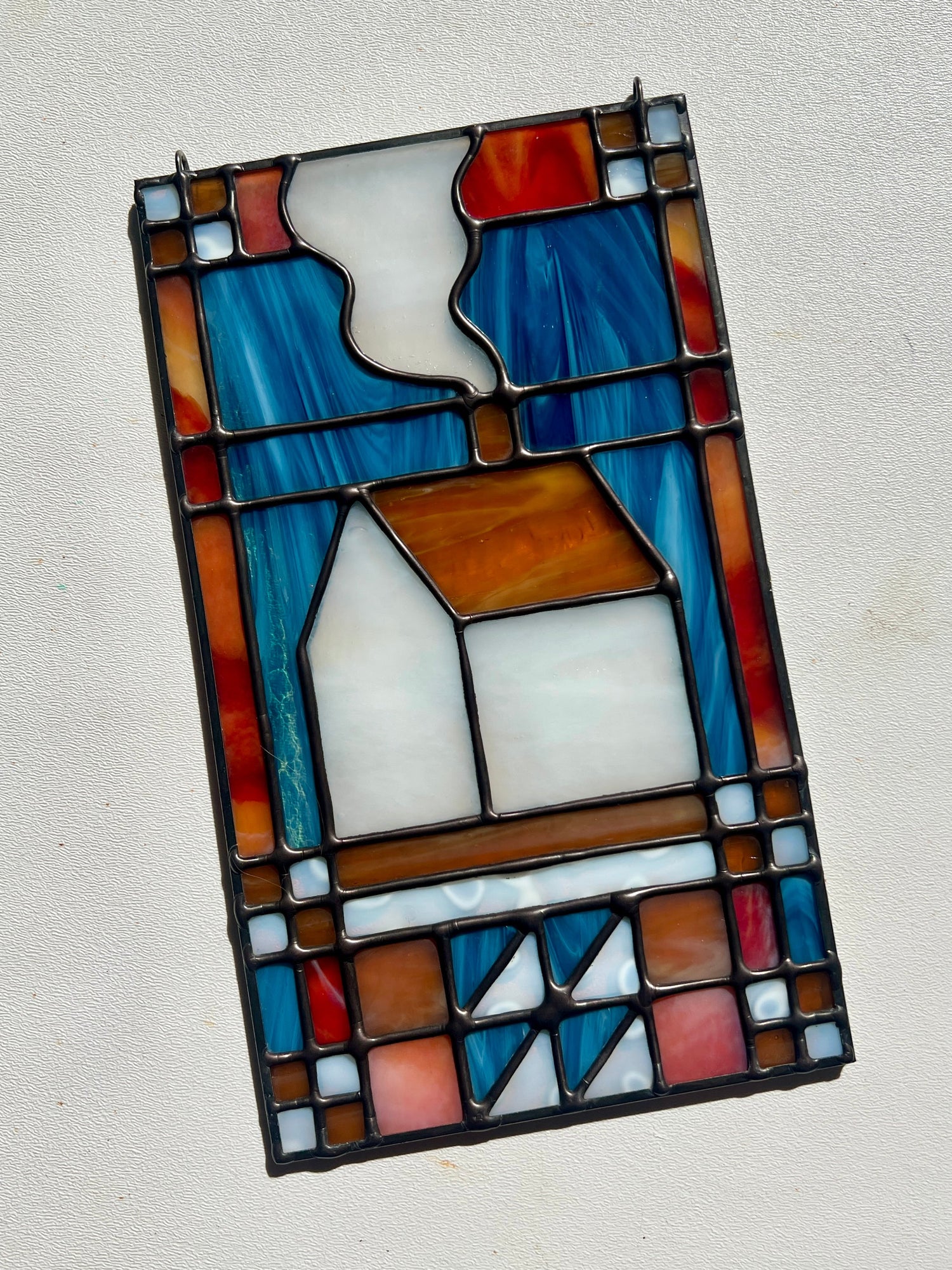 Stained glass textile inspired panel depicting a home in the center in shades of red, blue, white, and amber, resting on a white surface