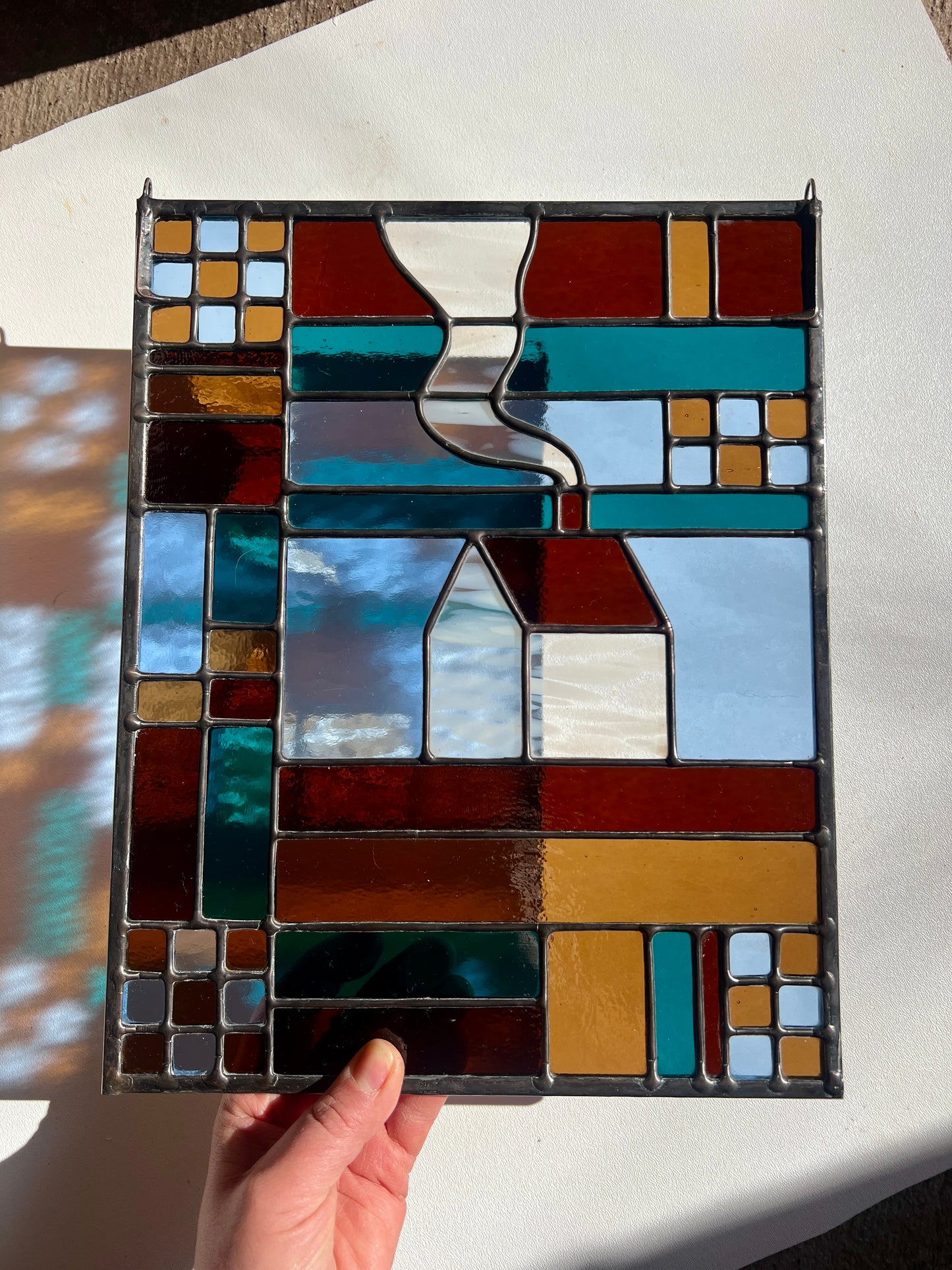 Stained glass textile inspired panel depicting a home in the center in shades of teal, brown, and blue, held by a hand in front of a white background with bright sunlight casting colorful refractions
