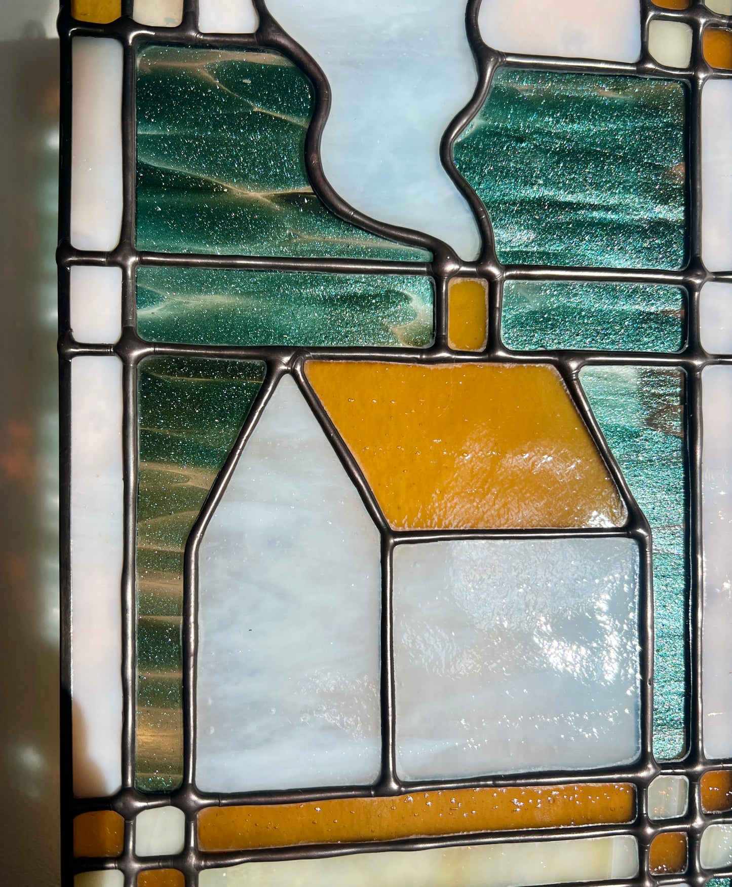 Close up of a textile inspired stained glass panel with a home in the center highlight sparkly teal and rick amber glass and smooth solder lines.