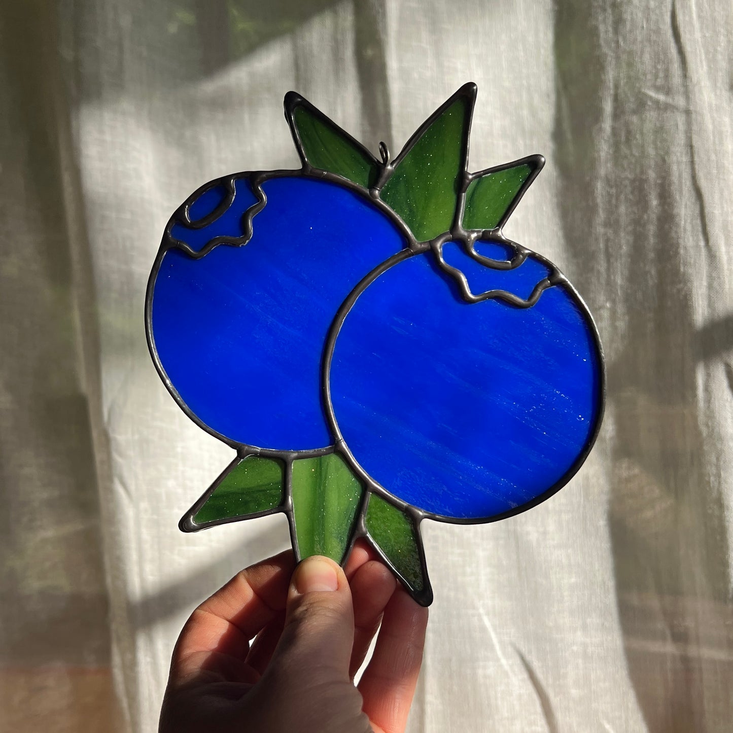 hand holding a stained glass suncatcher depicting a small bunch of blueberries with leaves in front of a window