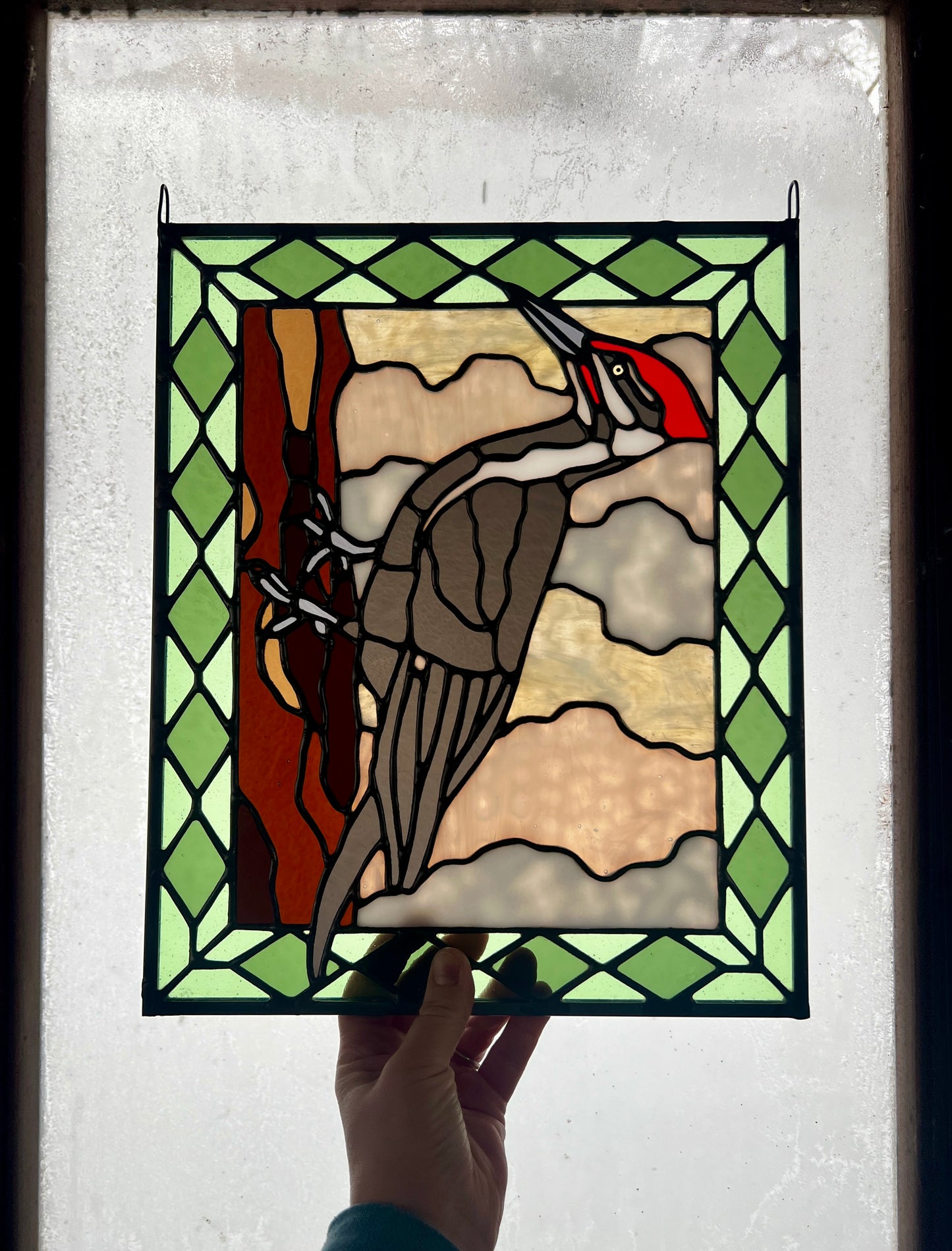 stained glass panel depicting a pileated woodpecker with a green diamond pattern border, held up in someone's hand