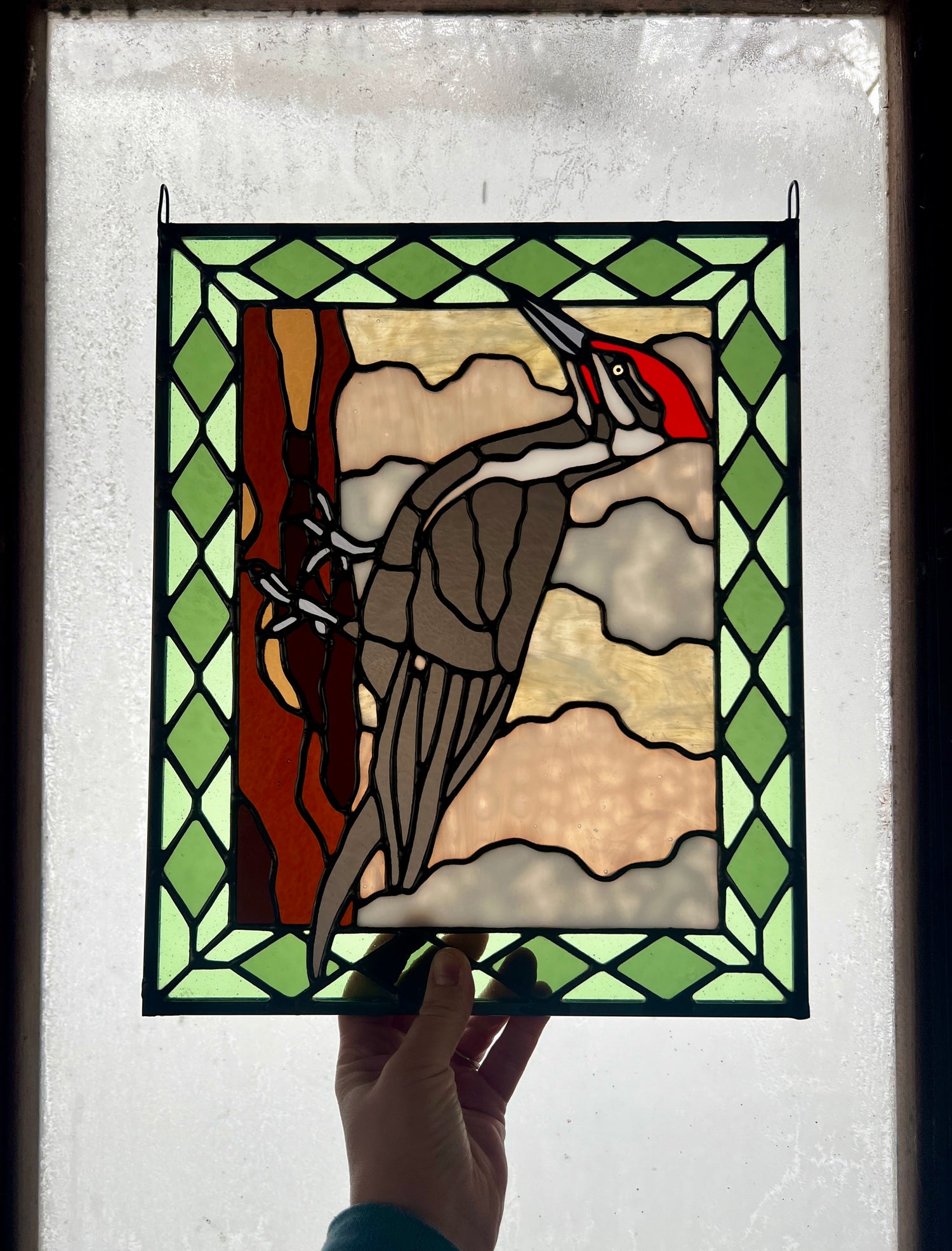 stained glass panel depicting a pileated woodpecker with a green diamond pattern border, held up in someone's hand