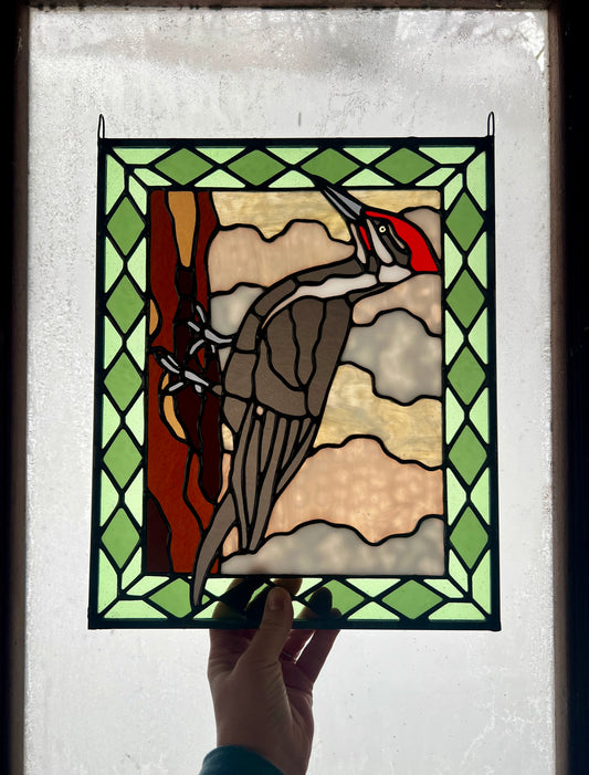 stained glass panel depicting a pileated woodpecker with a green diamond pattern border, held up in someone's hand