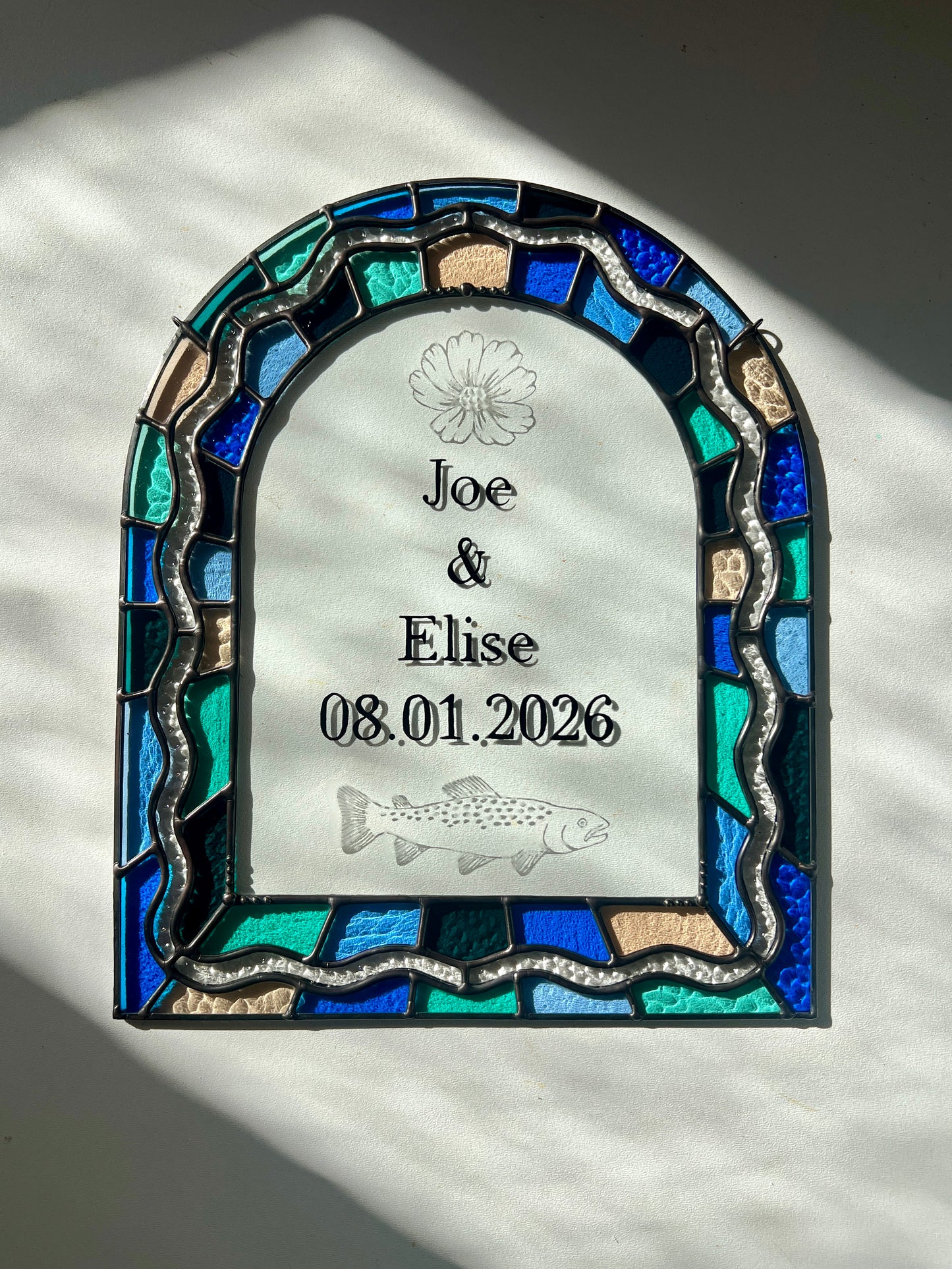 Stained glass panel in the shape of an arch with a multicolor blue, teal, turquoise, and pink border. Within the border is squiggle detail in clear textured glass. The center of the arch is clear uncolored glass, with personalized text and hand etched details of a cosmos flower and trout. The piece is resting against a white background with bright sunlight falling across it.