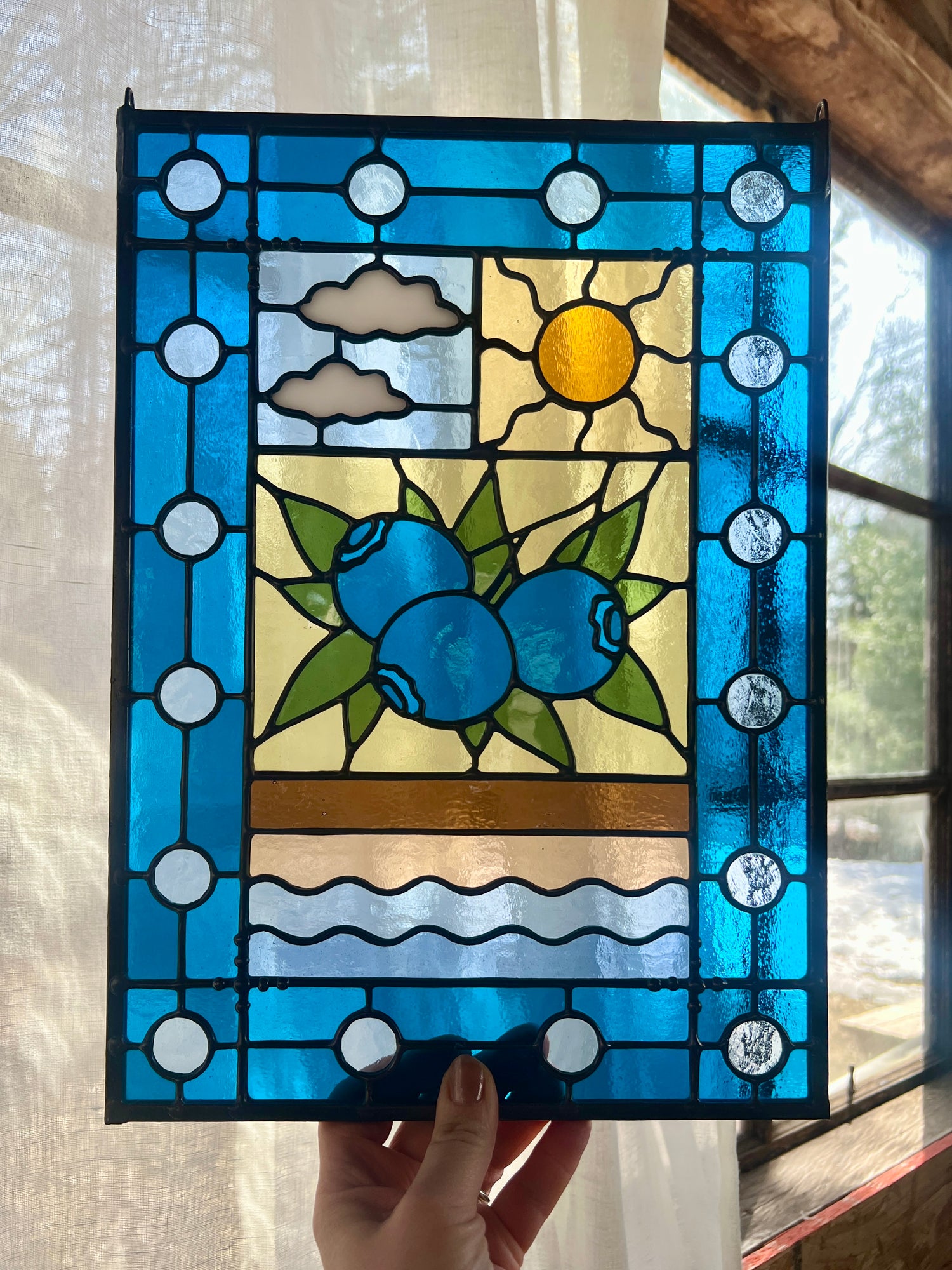stained glass panel with a simple compartment style design featuring clouds, sun, water, and a small bunch of blueberries in the middle with a blue border, held by a hand in front of a window