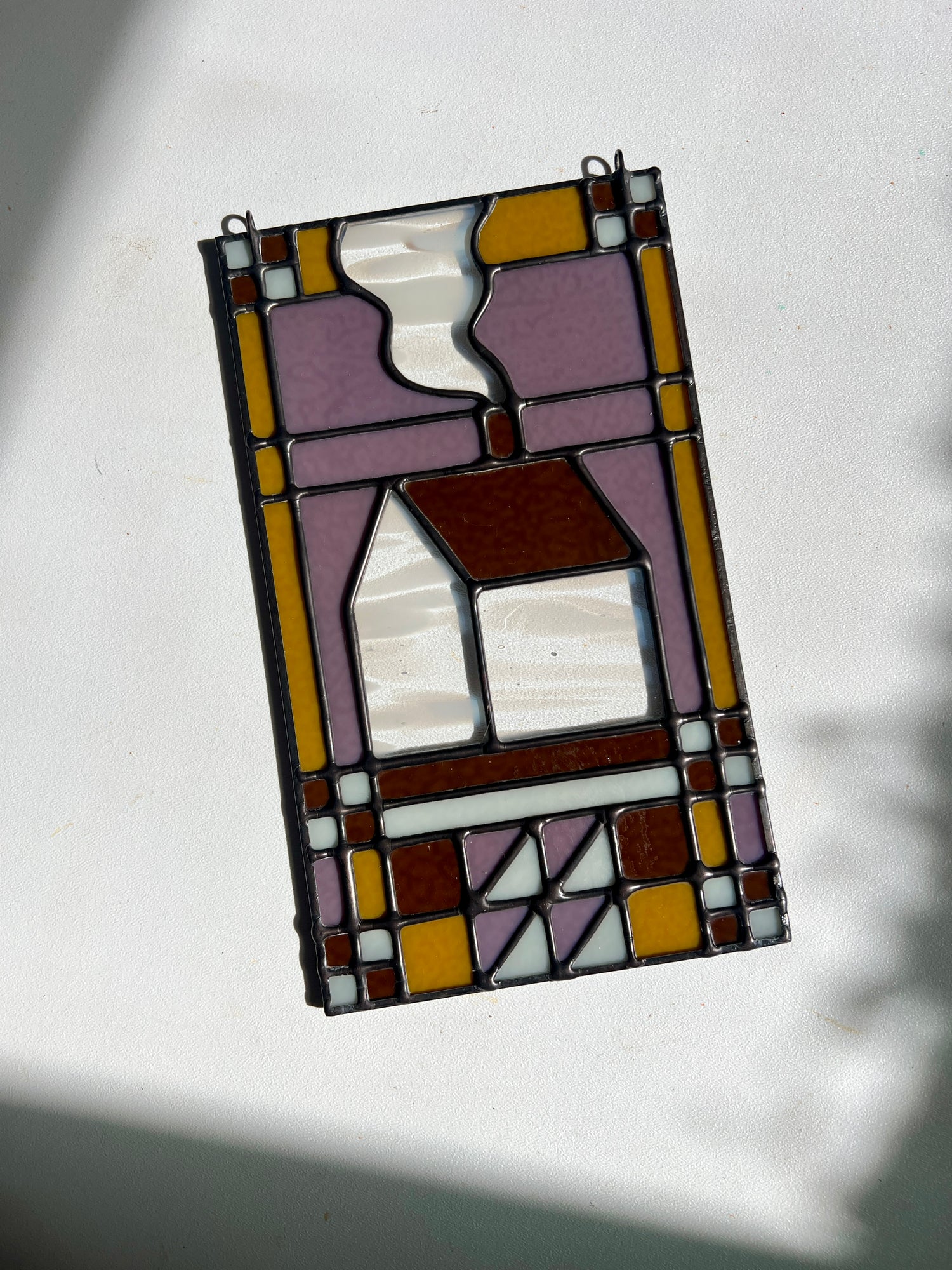 Stained glass textile inspired panel depicting a home in the center in shades of purple, white, brown, and bronze, set on a white surface with bright sunlight cast across