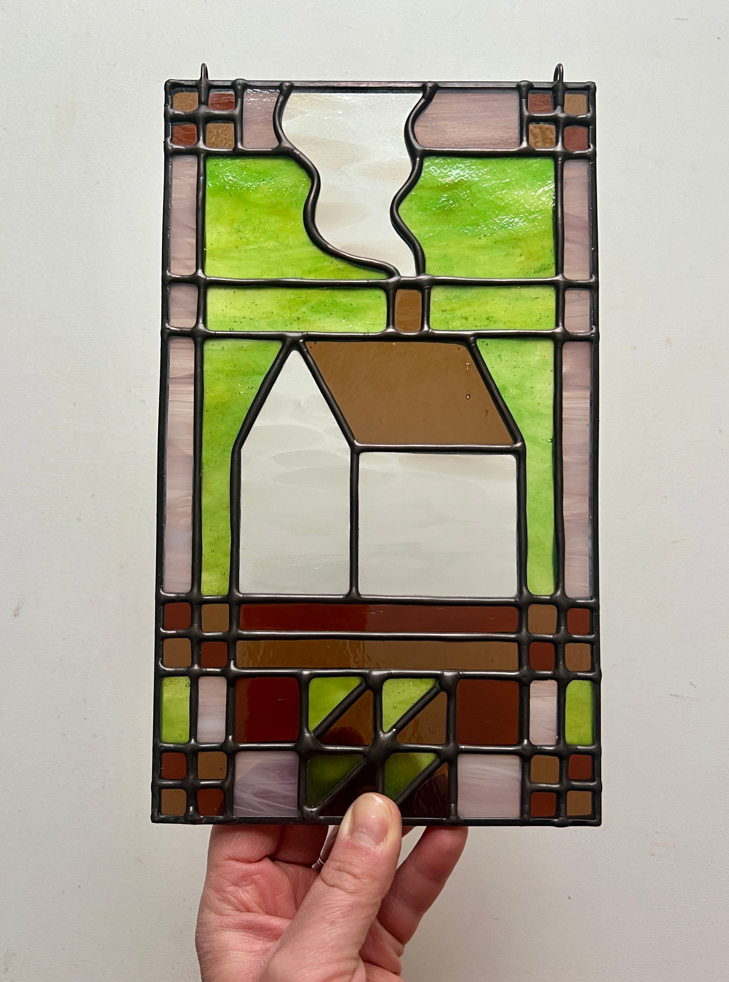 Stained glass textile inspired panel depicting a home in the center in shades of green, pink, white, and brown, held by a hand against a white background